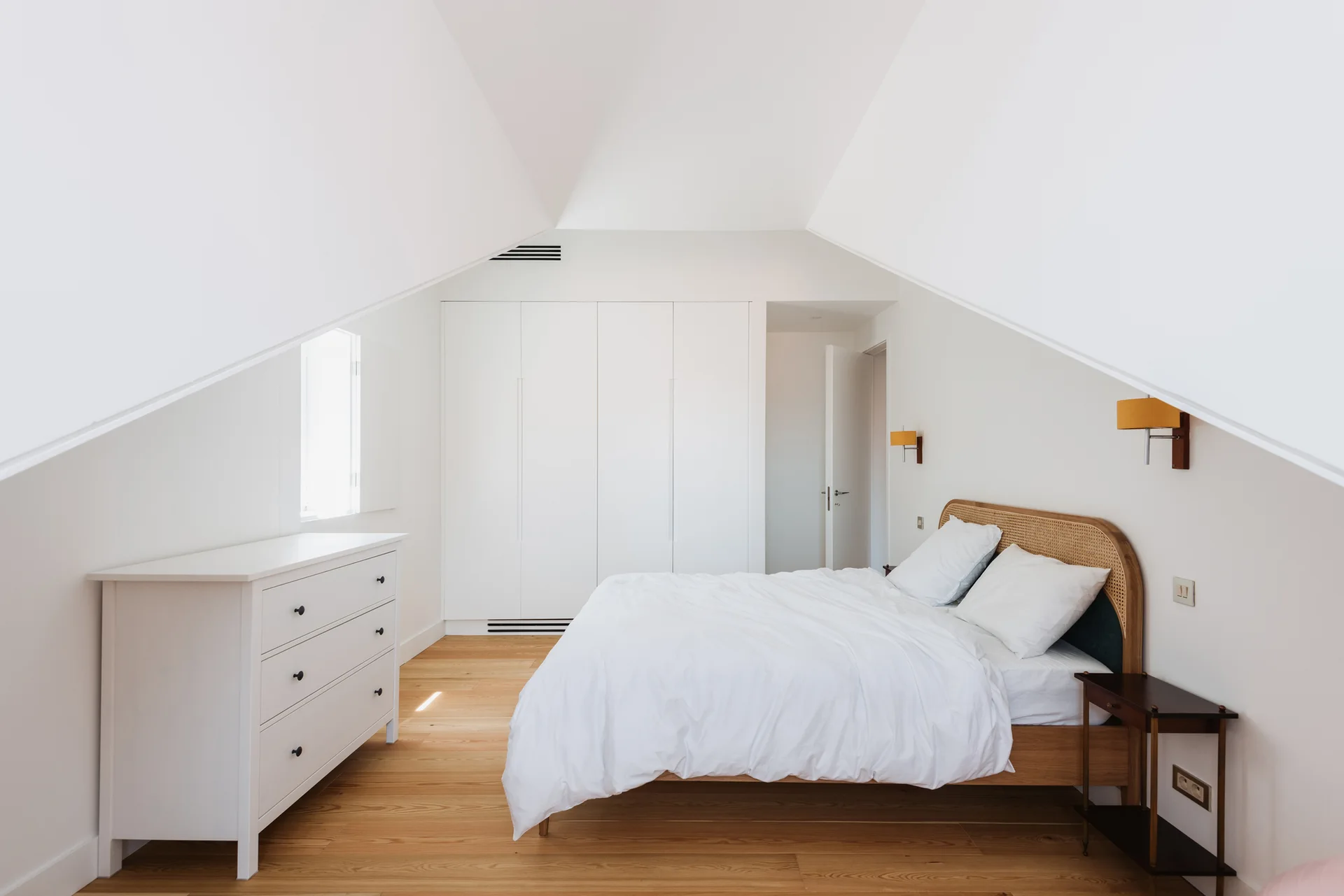 A minimalist attic bedroom features a wood bed with a rattan headboard and white built-in wardrobes beneath a pitched ceiling.