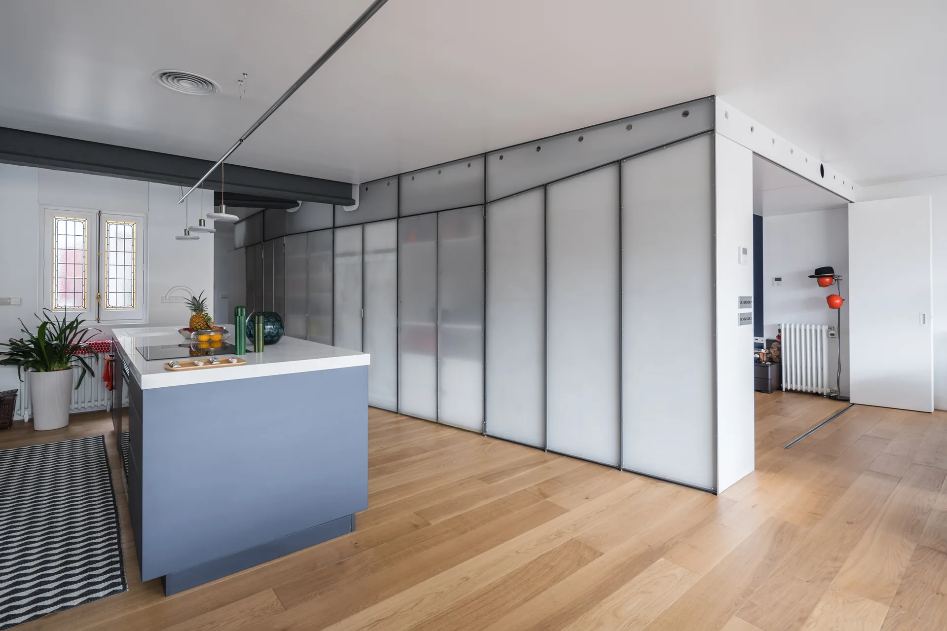 A modern open-plan kitchen features a light blue kitchen island, natural wood flooring, and a large sliding partition made of frosted glass panels.