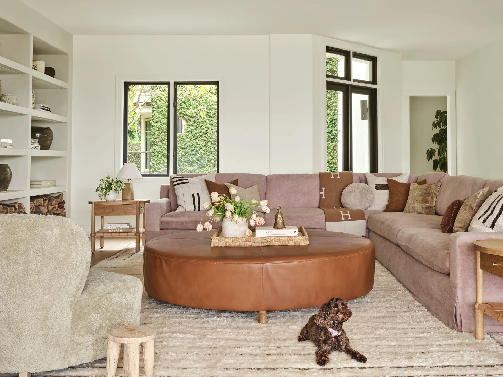 A bright living room features a mauve sectional sofa, a large round brown leather ottoman, and built-in shelving beside windows.