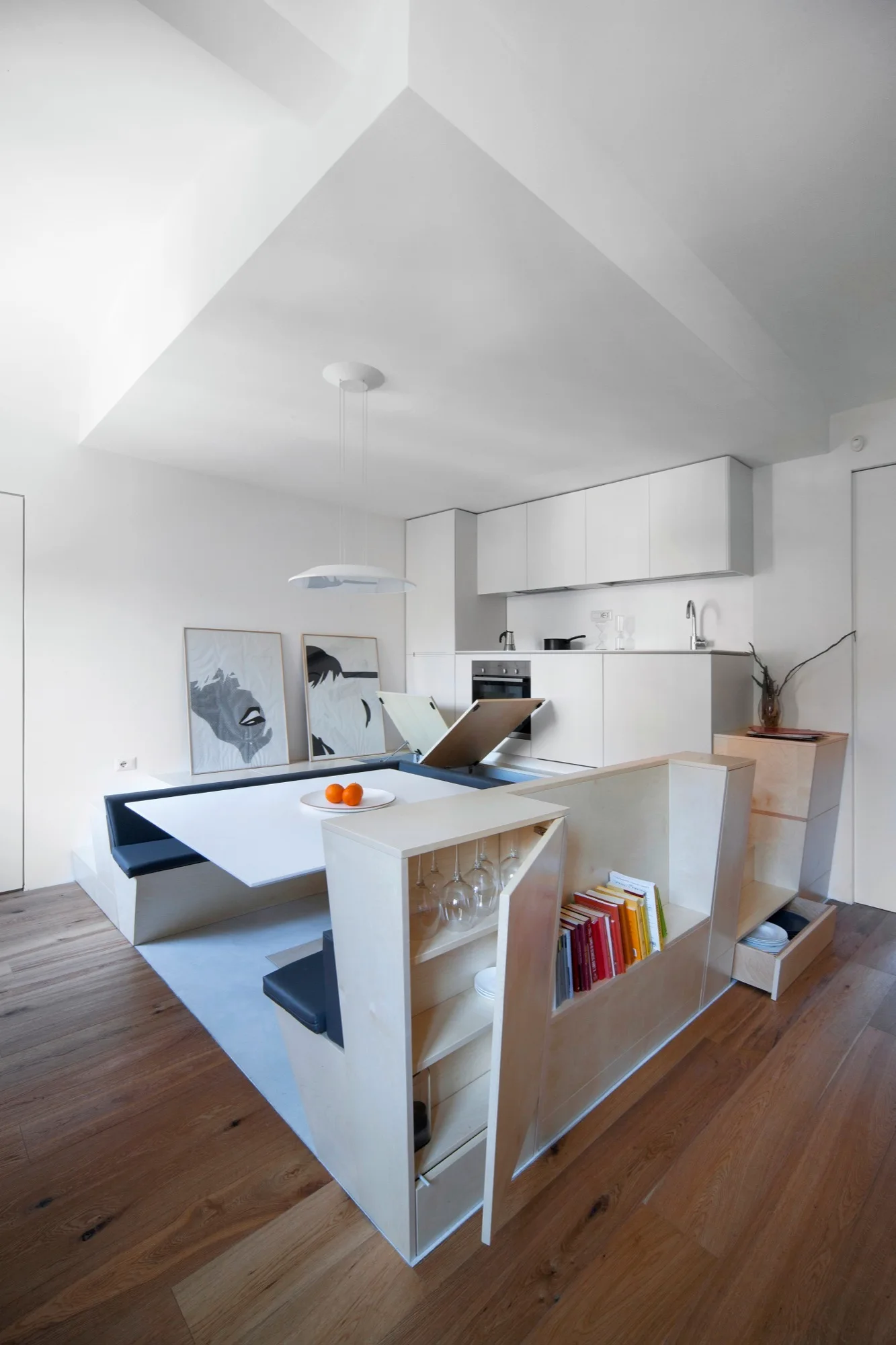 A modern kitchen features a custom plywood dining bench with integrated bookshelves, storage drawers, and a white minimalist table.