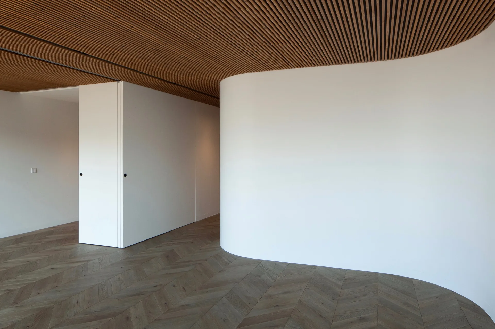 A minimalist interior hallway features a distinctive curved white wall, a linear wood slatted ceiling, and herringbone parquet wood flooring.