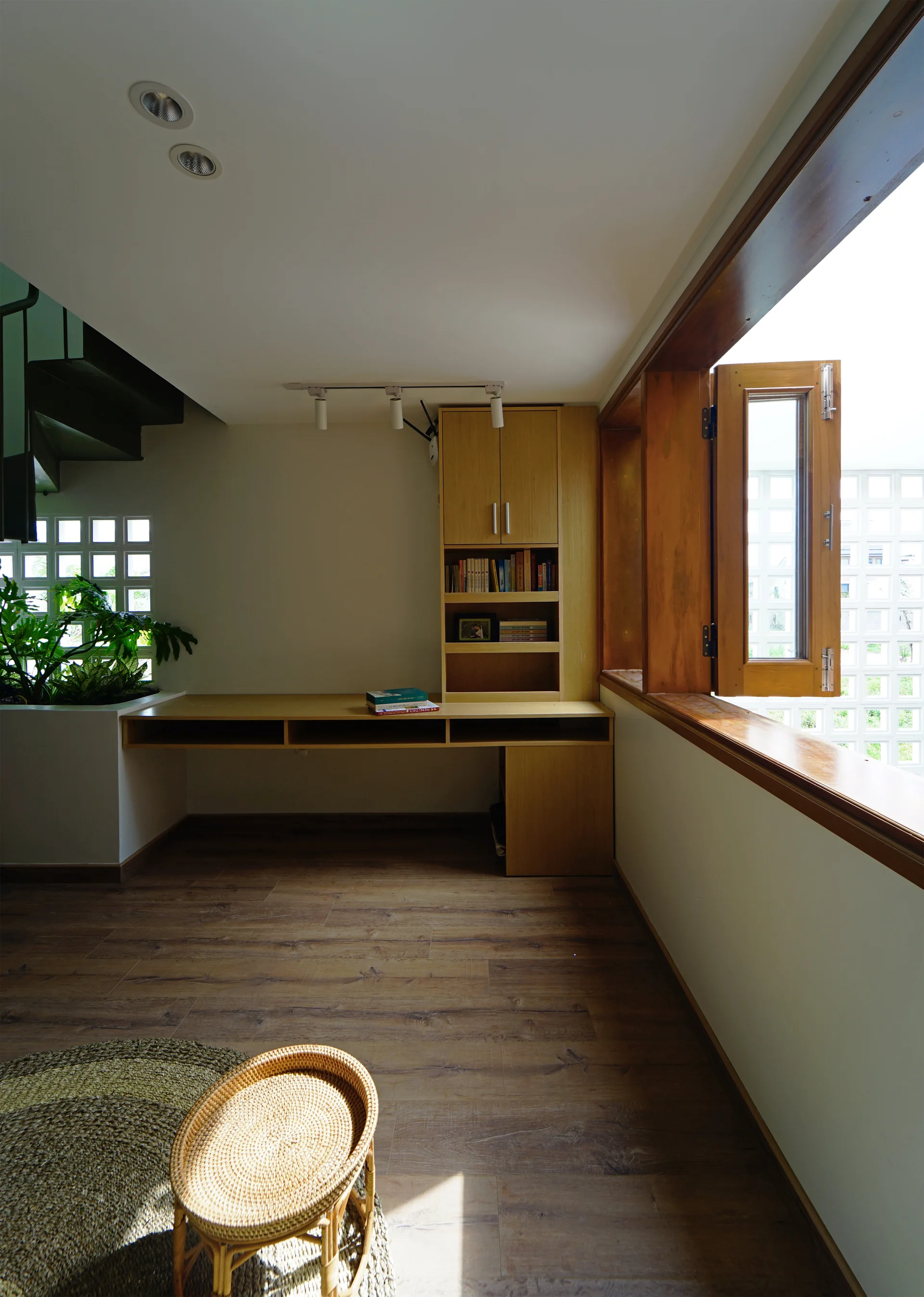 A minimalist home office features a light wood built-in desk, matching tall shelving, a wide wooden window frame, and a corner plant container.