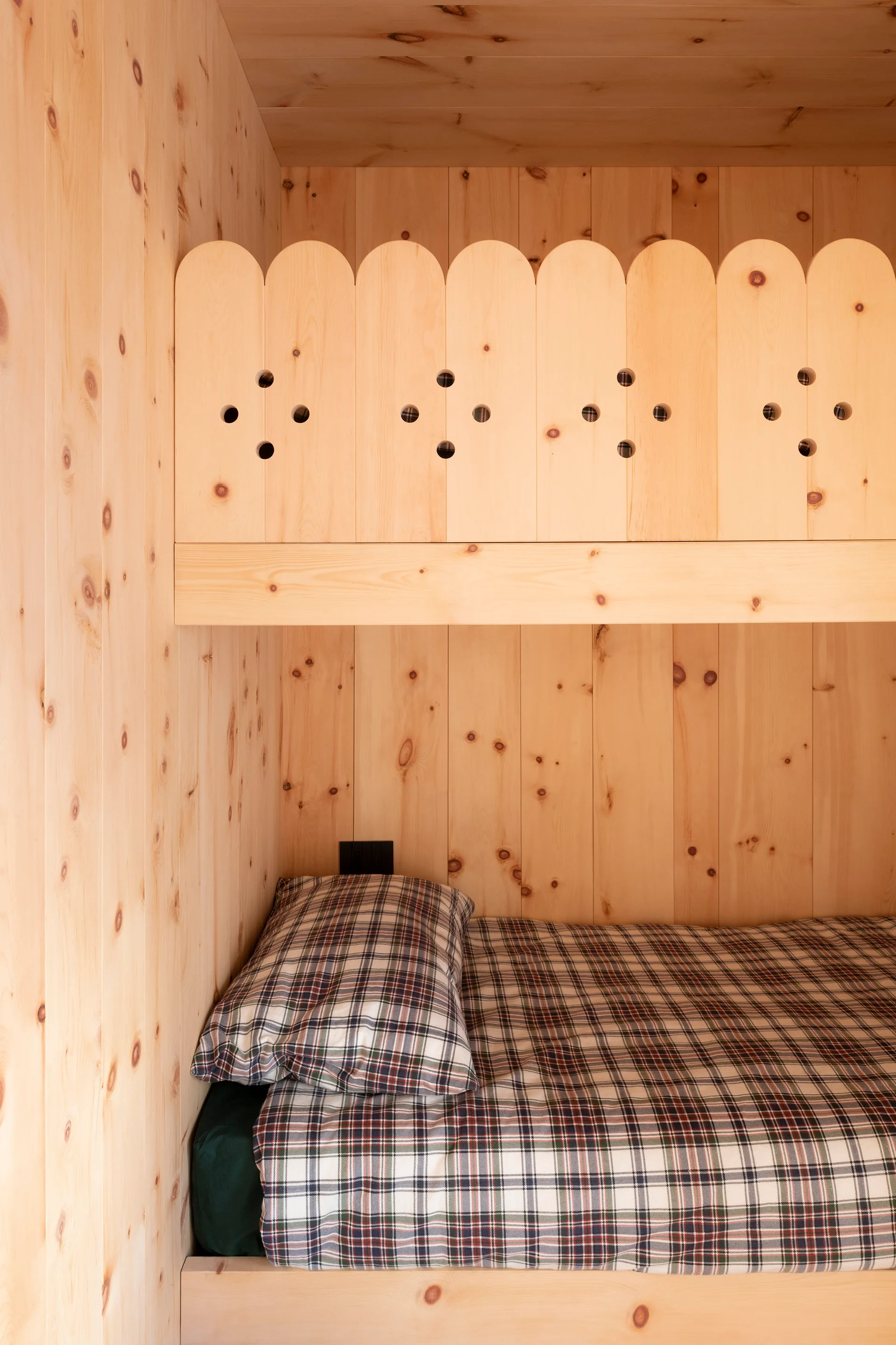 A built-in light wood bunk bed nook lined with knotty pine paneling, featuring a scalloped decorative guardrail and plaid bedding.