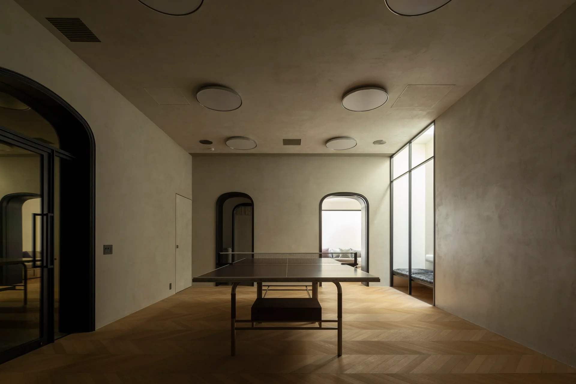 A minimalist recreation room featuring a central table tennis table, light oak herringbone flooring, and arched doorways set into neutral plaster walls.
