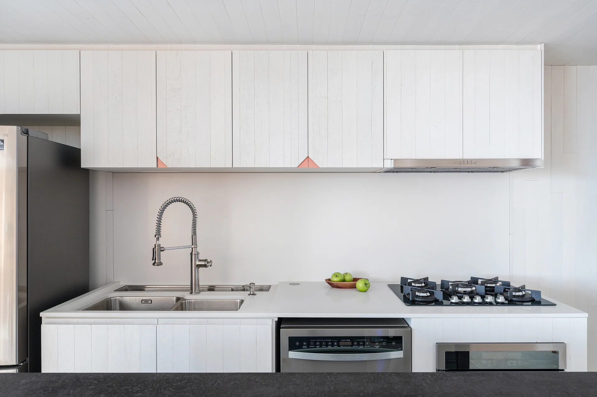 A modern kitchen featuring white vertical-slat cabinets with small copper corner details, a double sink, and built-in stainless steel appliances.