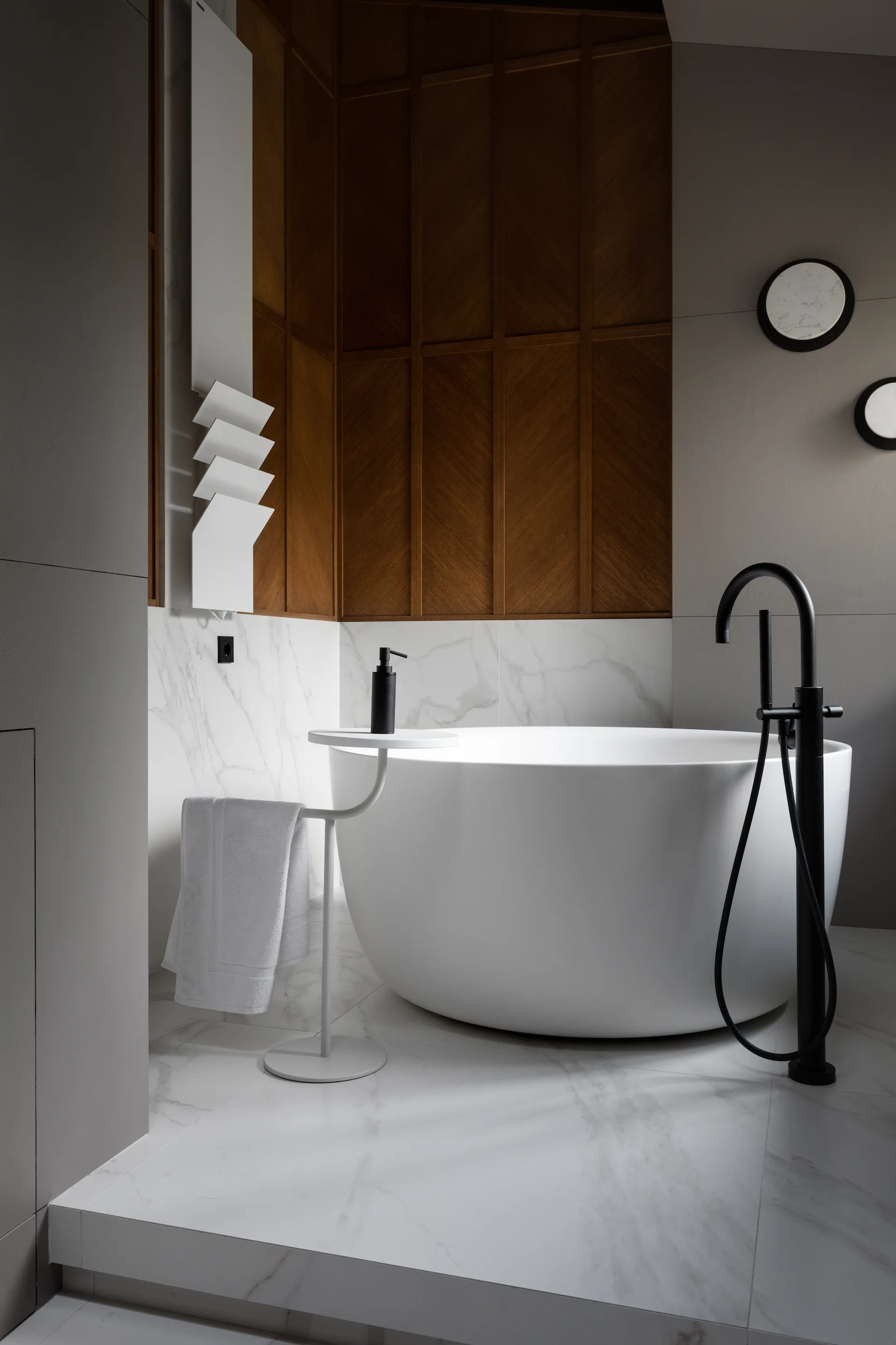 A white freestanding bathtub stands on a veined marble floor beside a wood herringbone accent wall and a matte black floor-mounted faucet.