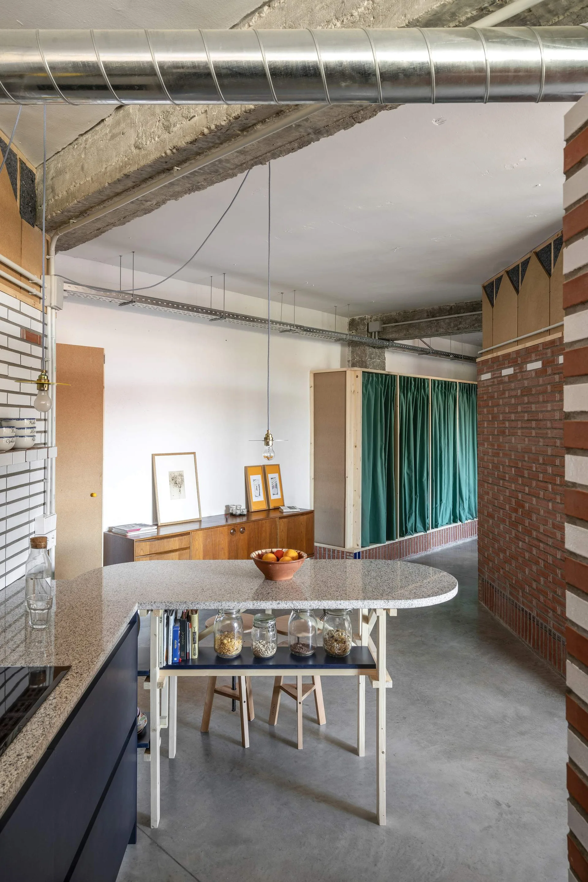 An industrial loft kitchen features a speckled stone island with wooden legs, exposed concrete beams, concrete flooring, and a red brick wall.