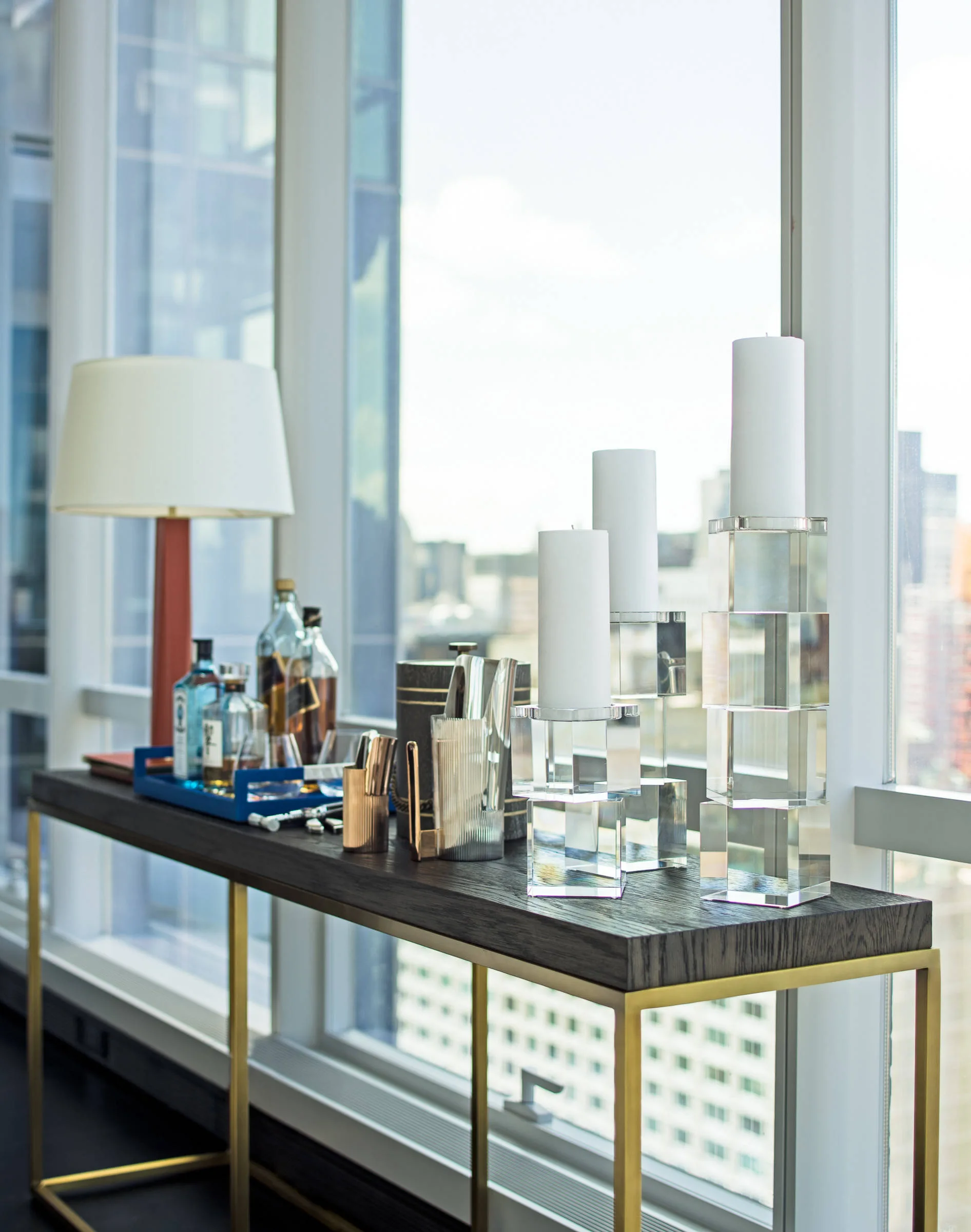 A dark wood bar console with a brass base holds liquor bottles, a lamp, and tall clear block candle holders in front of a window.