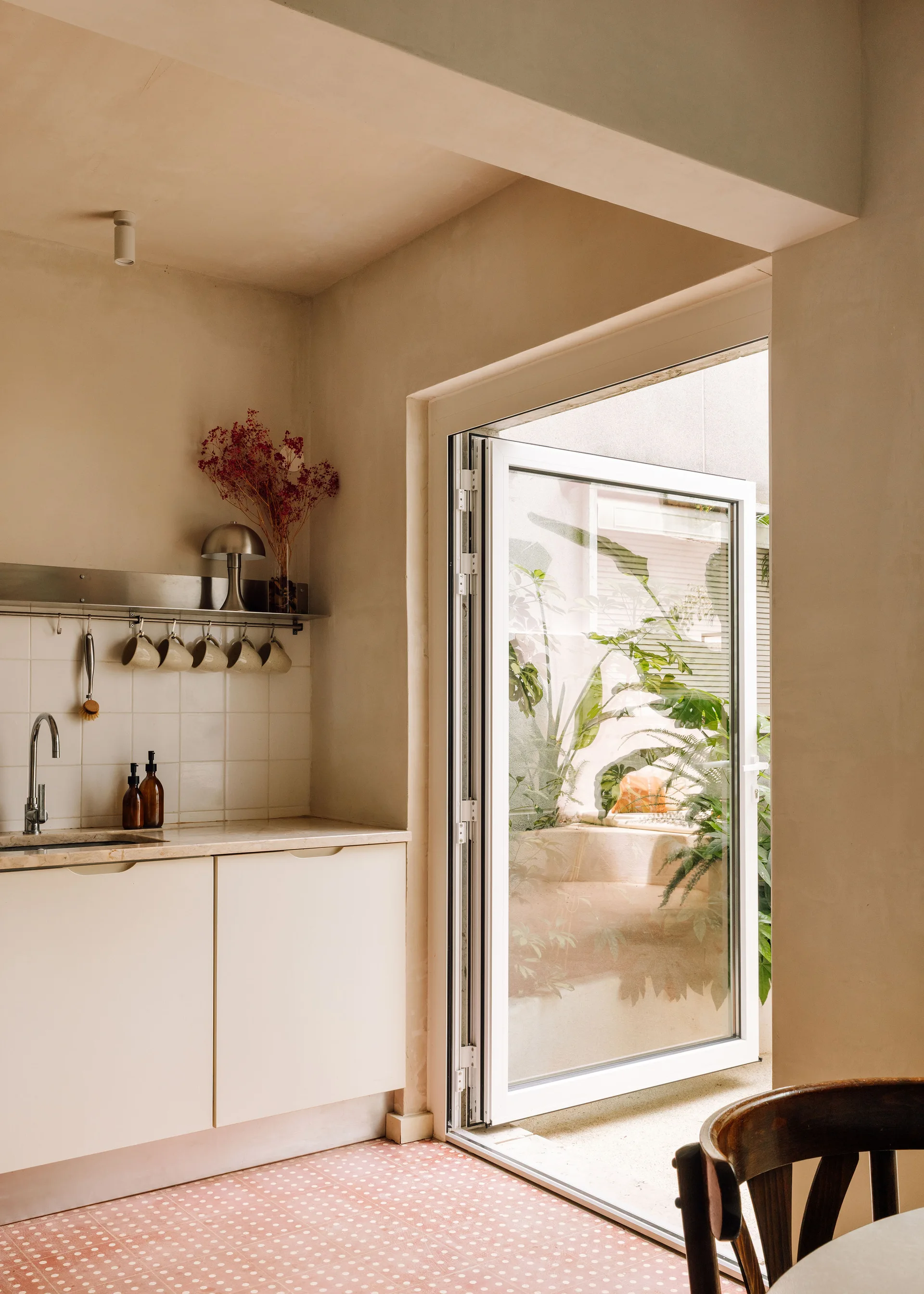 A warm beige kitchen featuring flat-front cabinets, a white tiled backsplash with a hanging mug rack, and a glass door opening to a green patio.