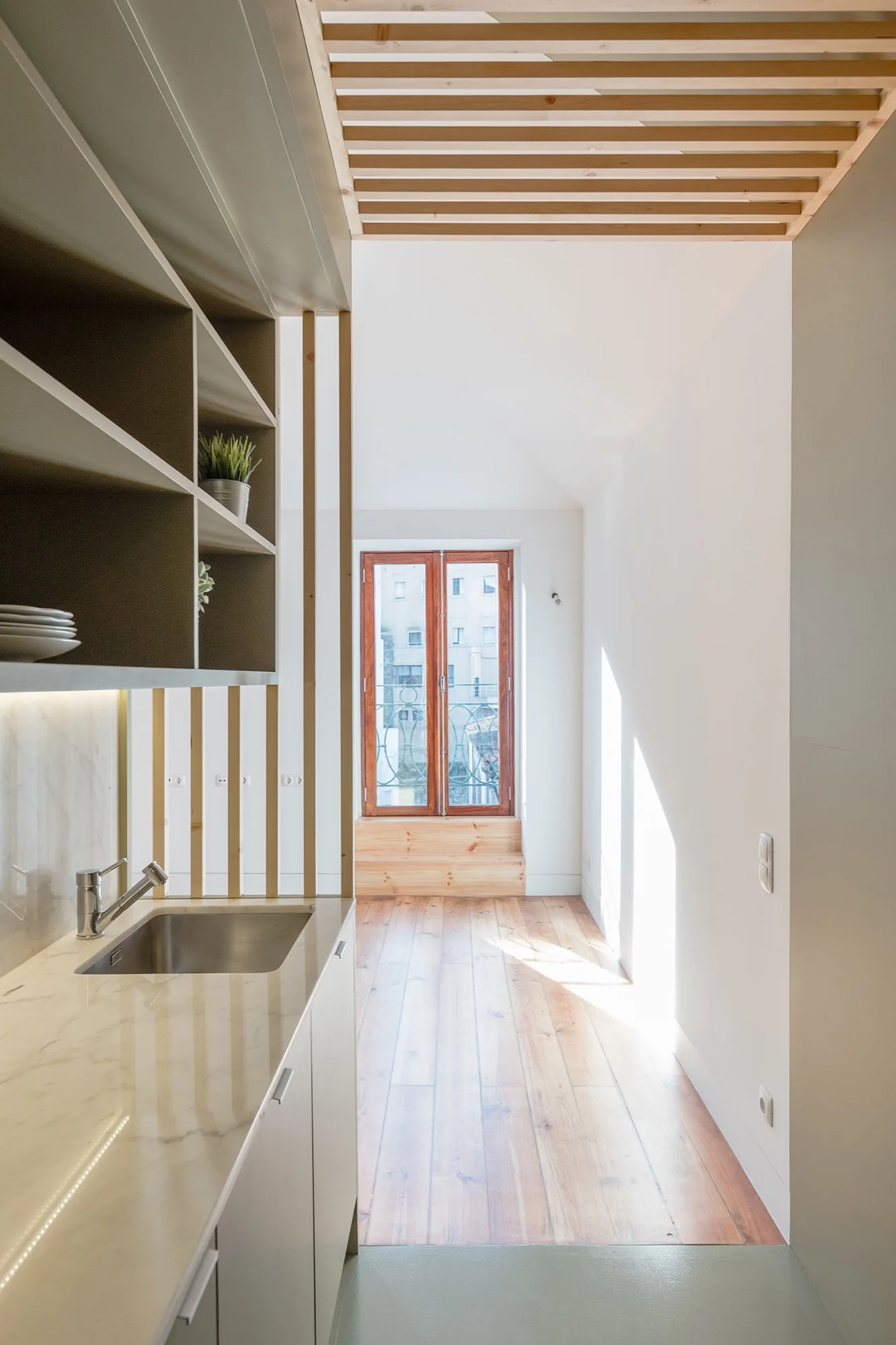 A narrow kitchen features a marble countertop with an integrated sink, matte grey shelving, and a vertical wood slat room divider next to wood floors.