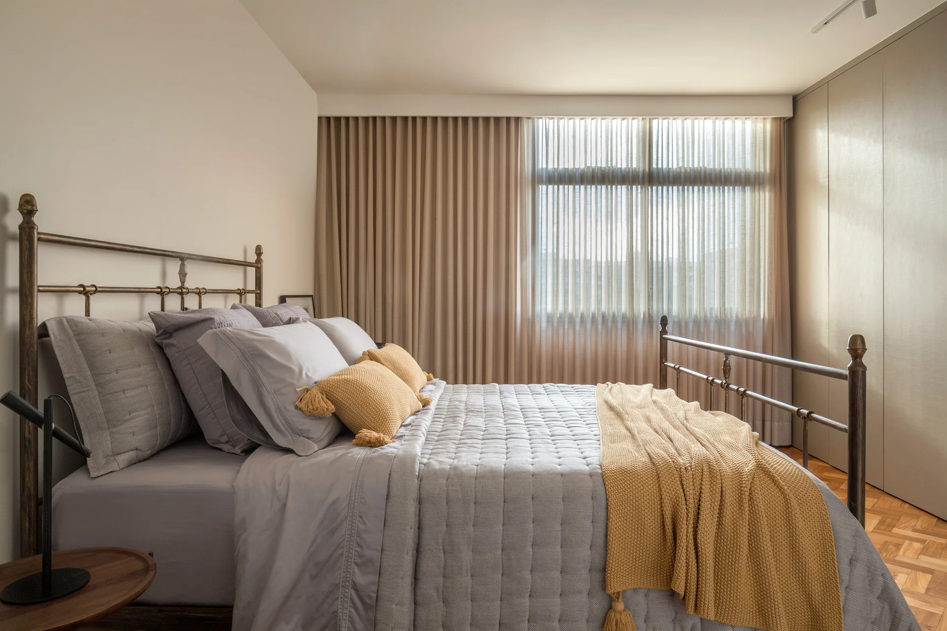 A bedroom featuring a metal frame bed with grey quilted bedding, mustard yellow accents, pleated curtains, and herringbone wood flooring.