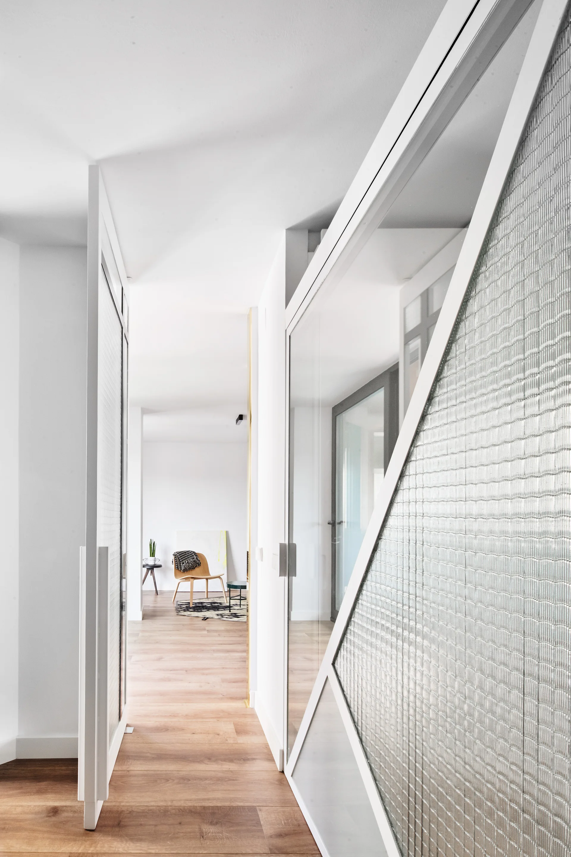A bright hallway featuring light wood flooring, clean white walls, and a large white-framed partition wall with textured ribbed glass panels.