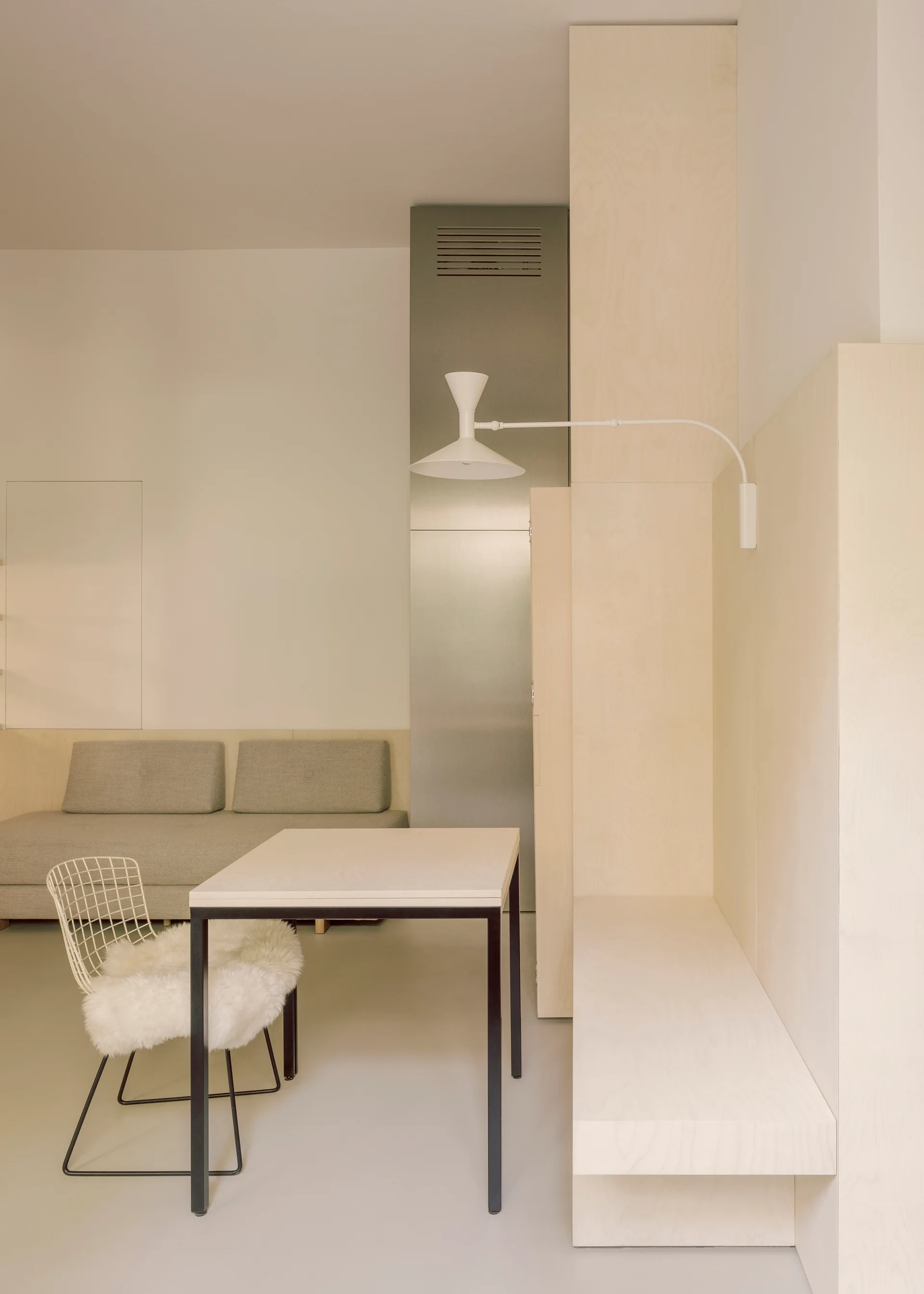 A minimalist dining area features light plywood wall paneling, an integrated bench, a square table with a wire chair, and a grey sofa.