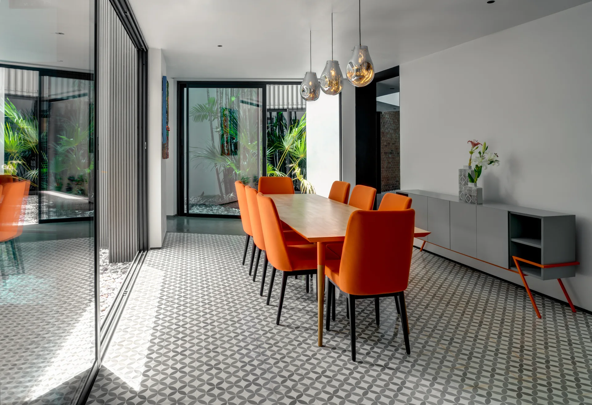 A modern dining room features a wooden table surrounded by orange chairs, a grey sideboard, and patterned floor tiles next to large glass sliding doors.