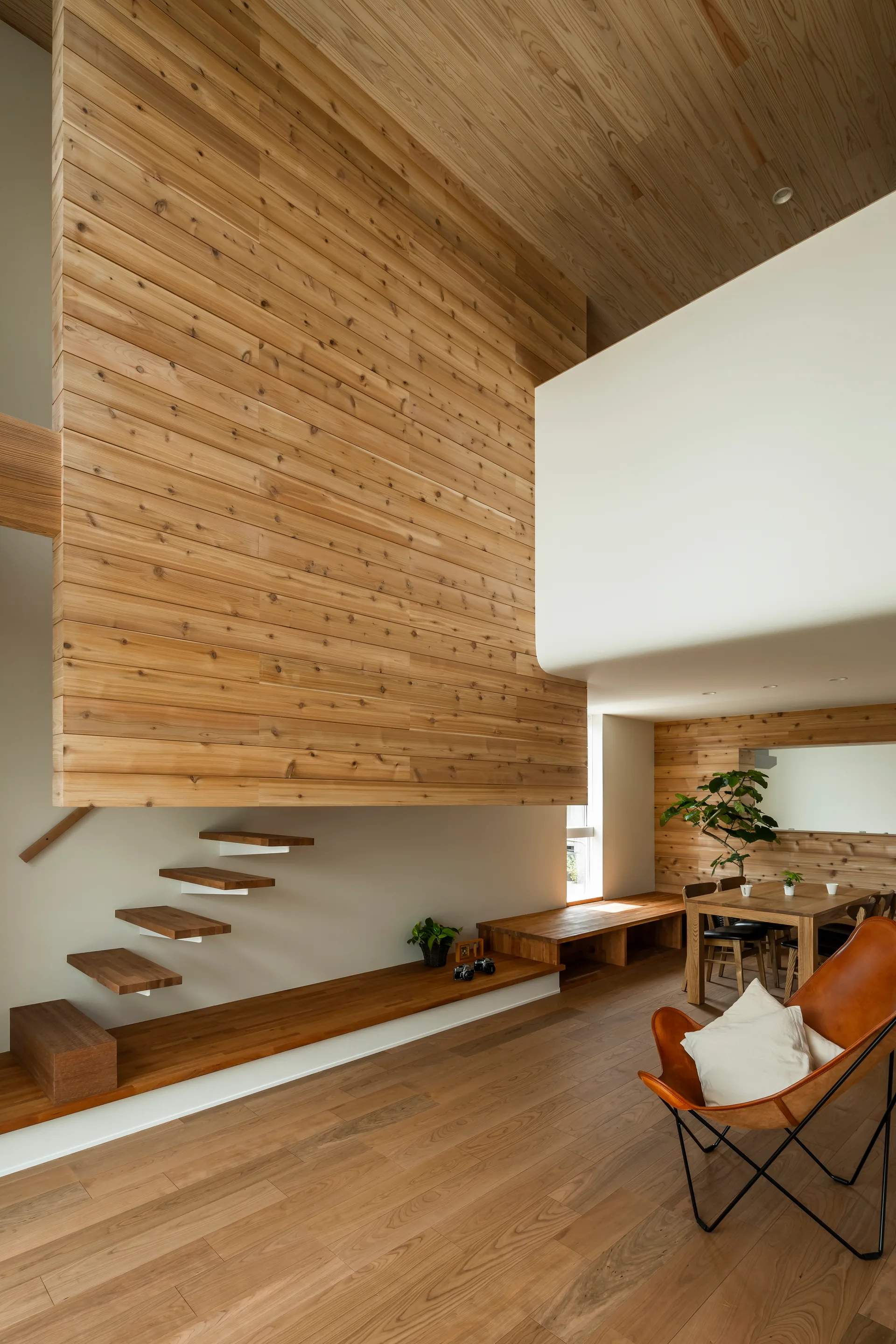 A modern open-concept living area featuring a floating wooden staircase, extensive wood wall paneling, and a leather butterfly chair in the foreground.