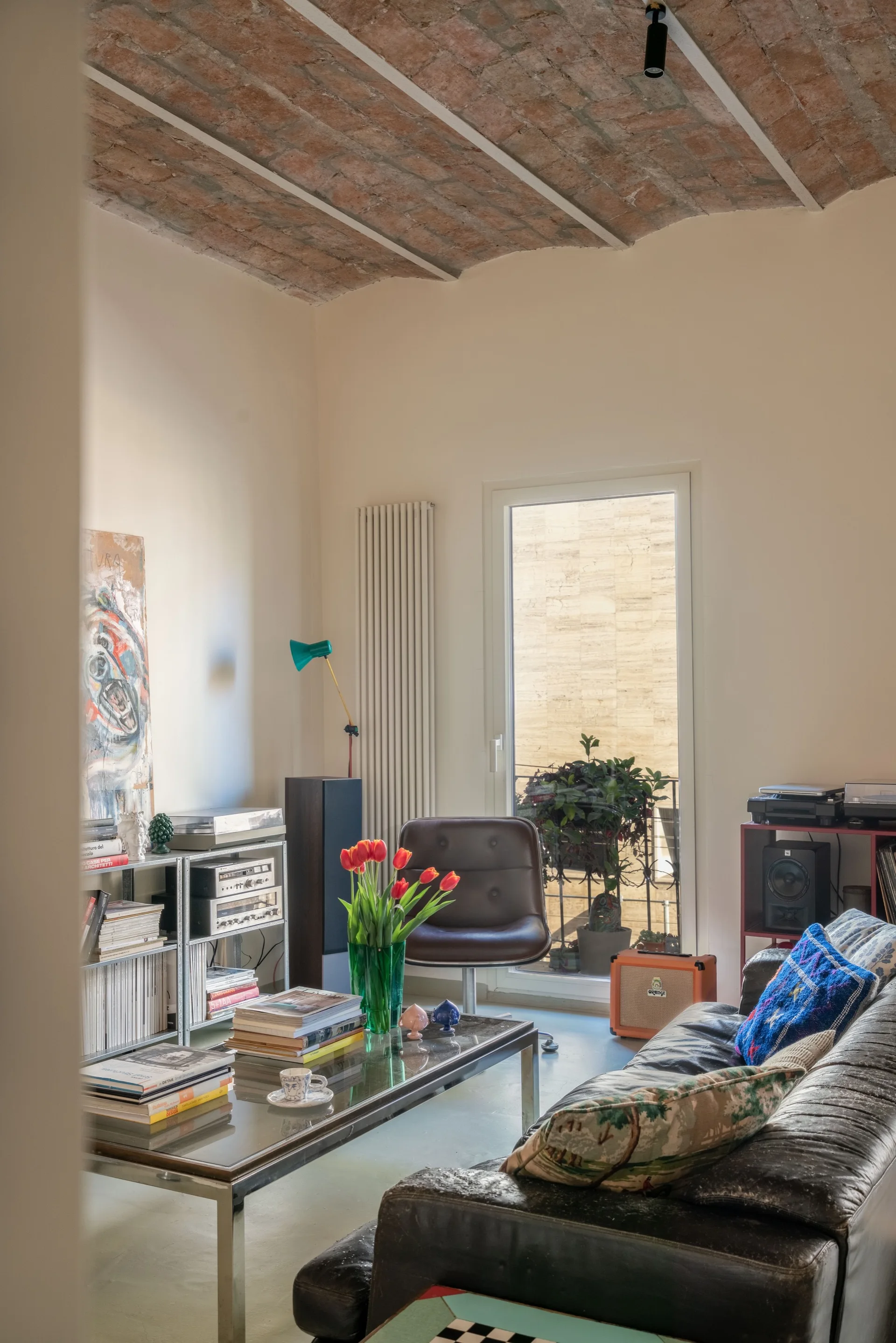 An eclectic living room featuring an exposed brick vaulted ceiling, a dark leather sofa, a glass coffee table, and a white metal shelving unit.