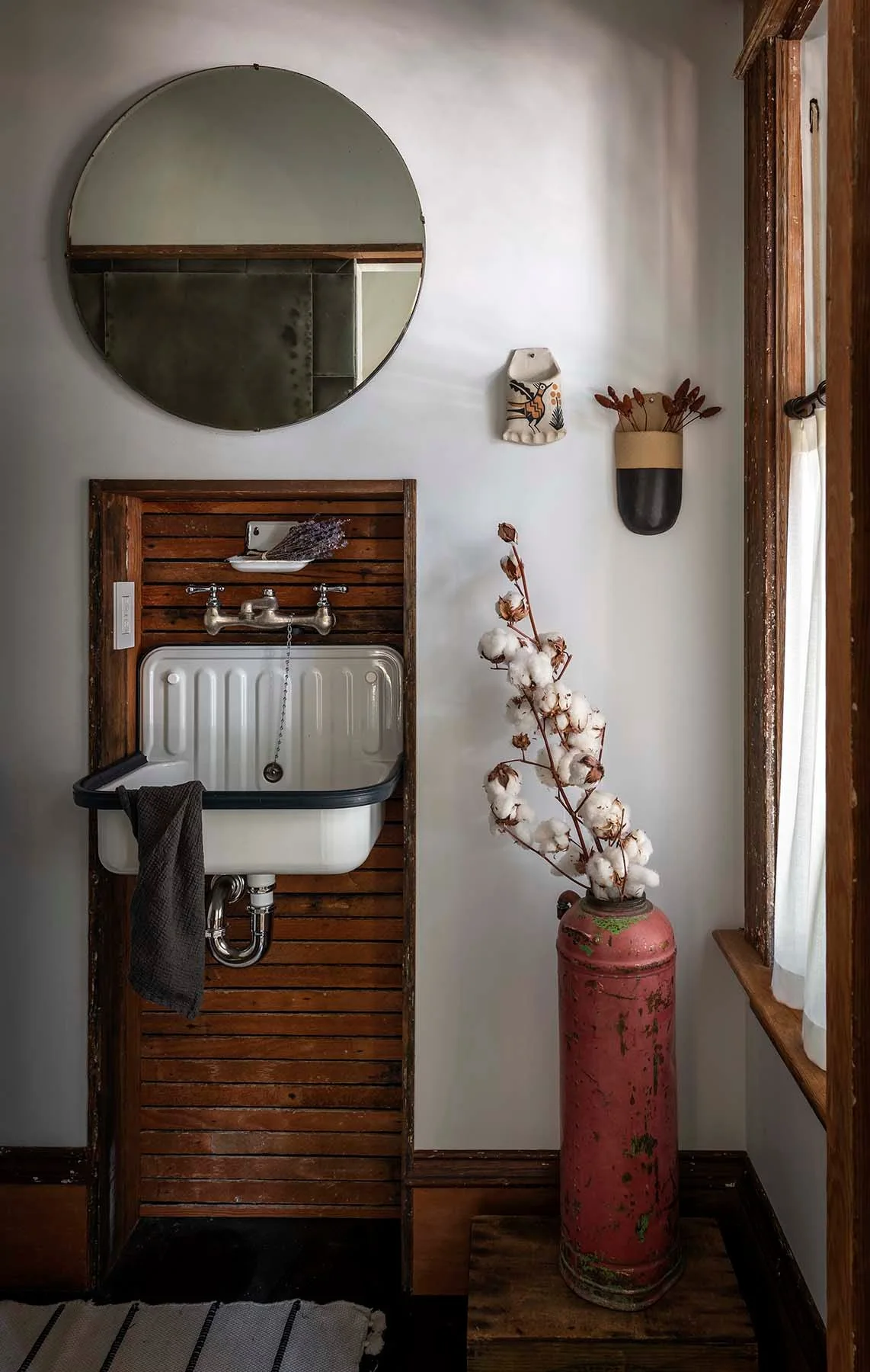 A white wall-mounted utility sink is set against horizontal wood slats, beneath a round mirror and next to a vintage red cylinder vase.