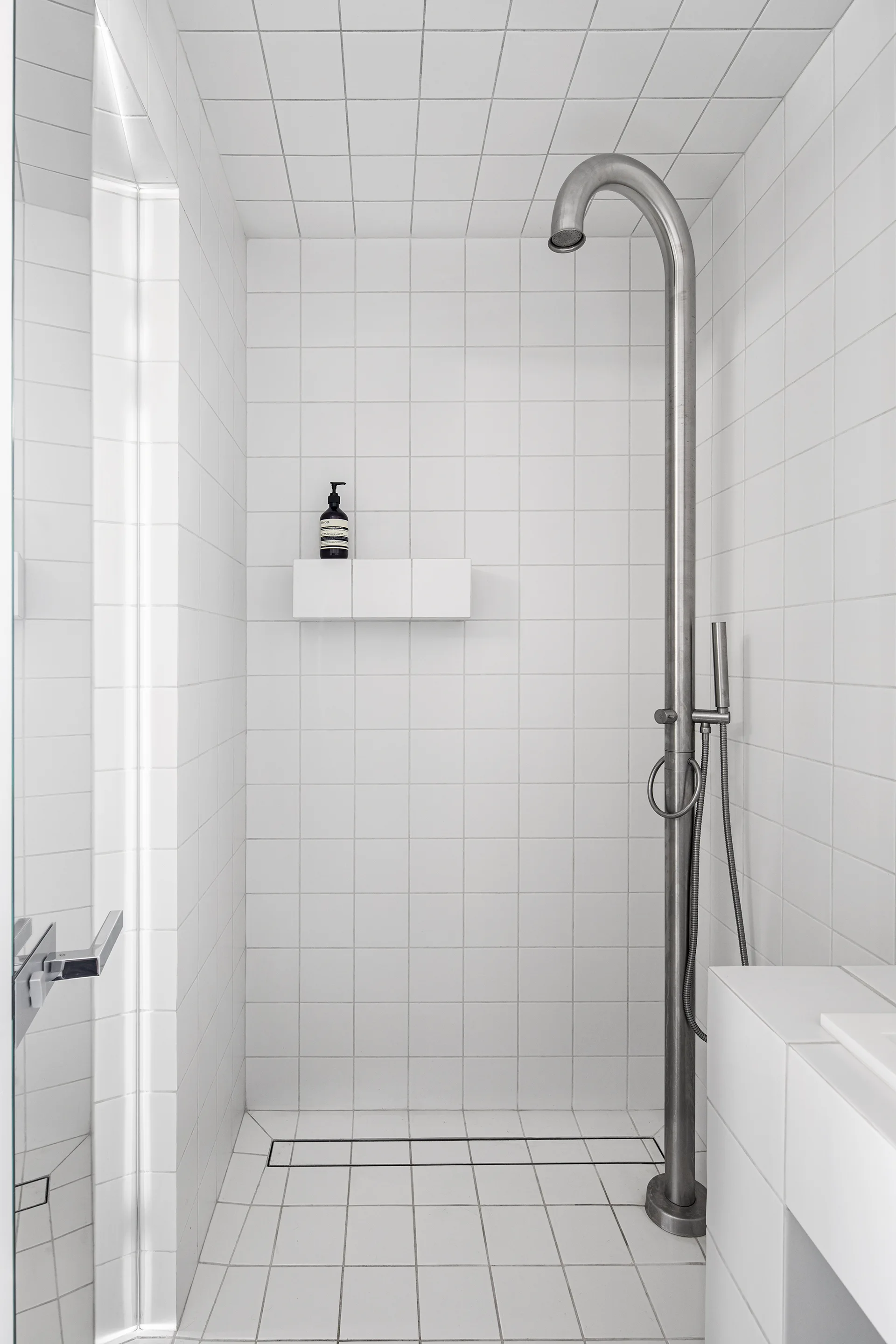 A minimalist shower stall clad in white square grid tiles featuring a tall curved stainless steel exposed pipe shower fixture.