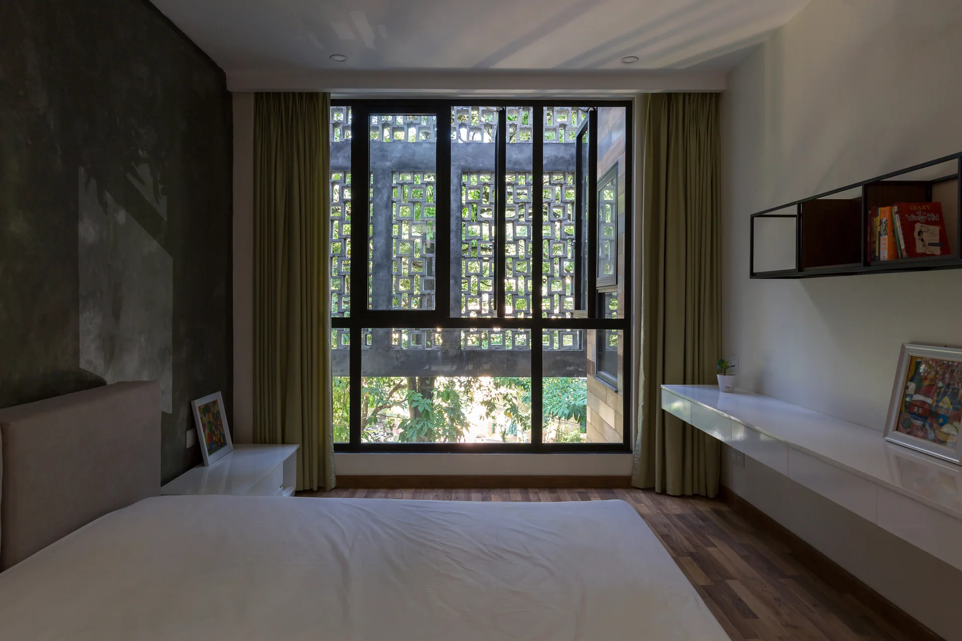 A minimalist bedroom features a large window with olive drapes looking out at a decorative concrete screen next to a white floating desk.