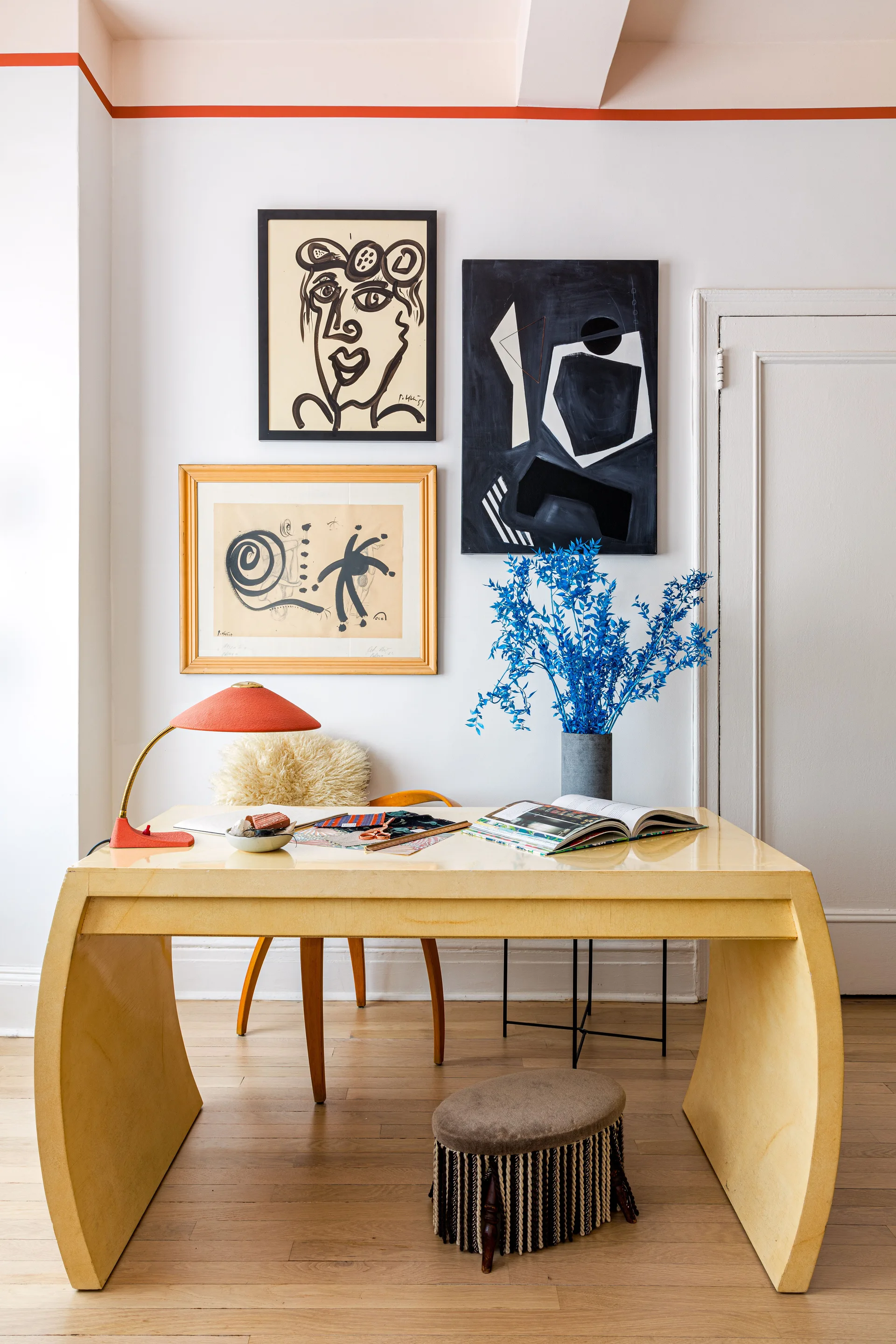 An eclectic home office features a pale yellow curved desk, abstract framed art, and an orange desk lamp.