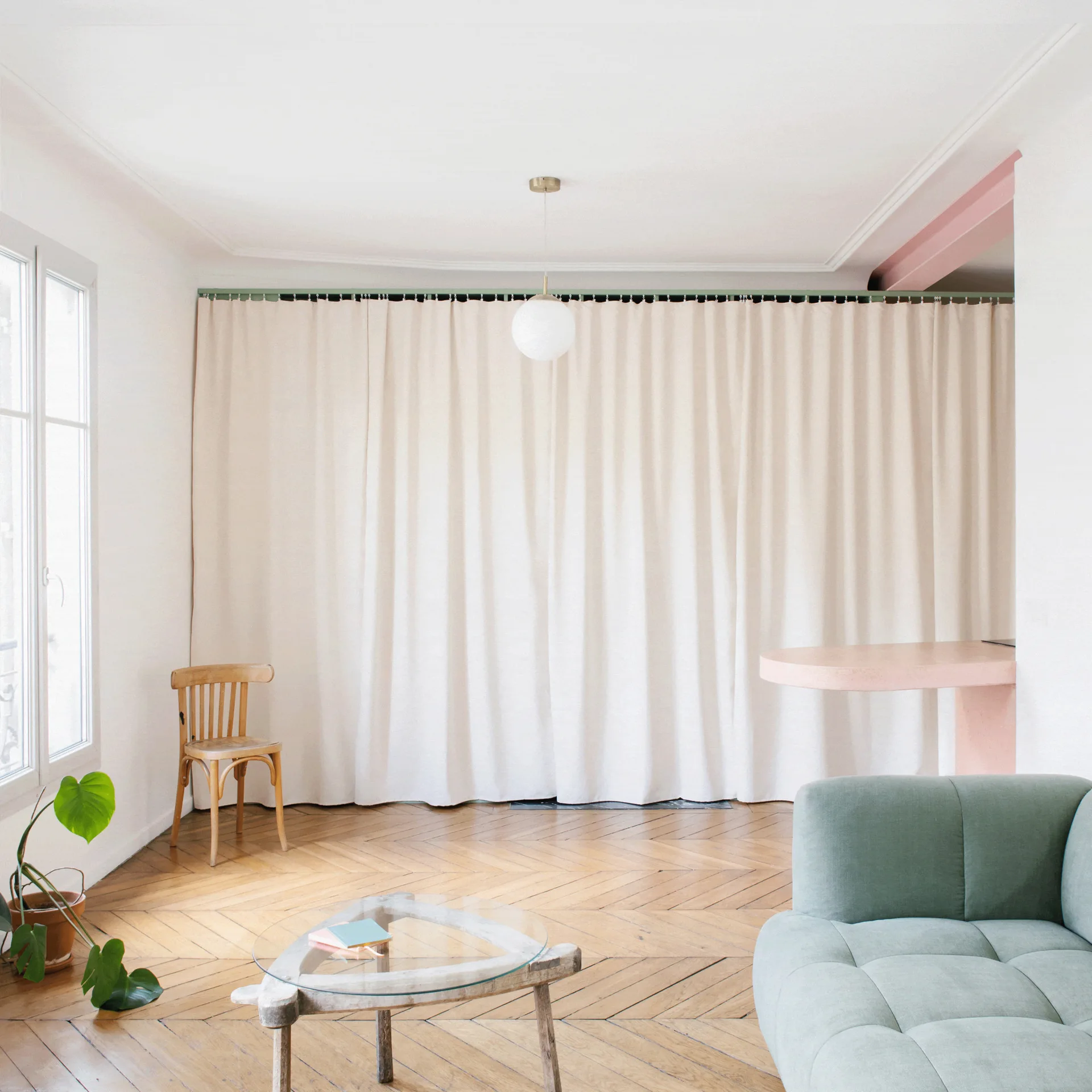 A minimalist living room features a mint green sofa, a glass coffee table, and a wooden chair on herringbone parquet flooring.