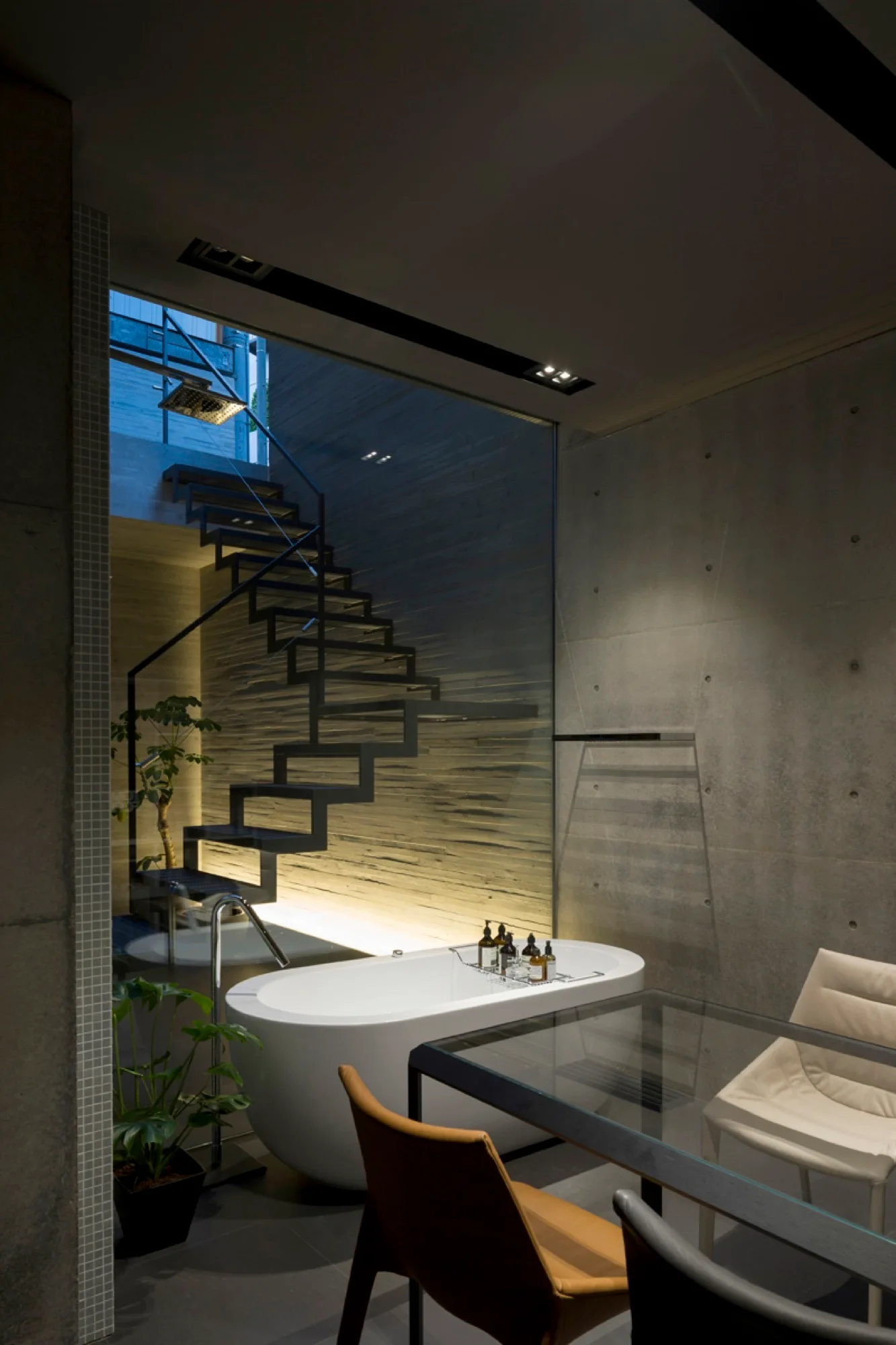A white freestanding bathtub sits near a concrete wall and a large glass partition showing a black floating staircase lit from below.
