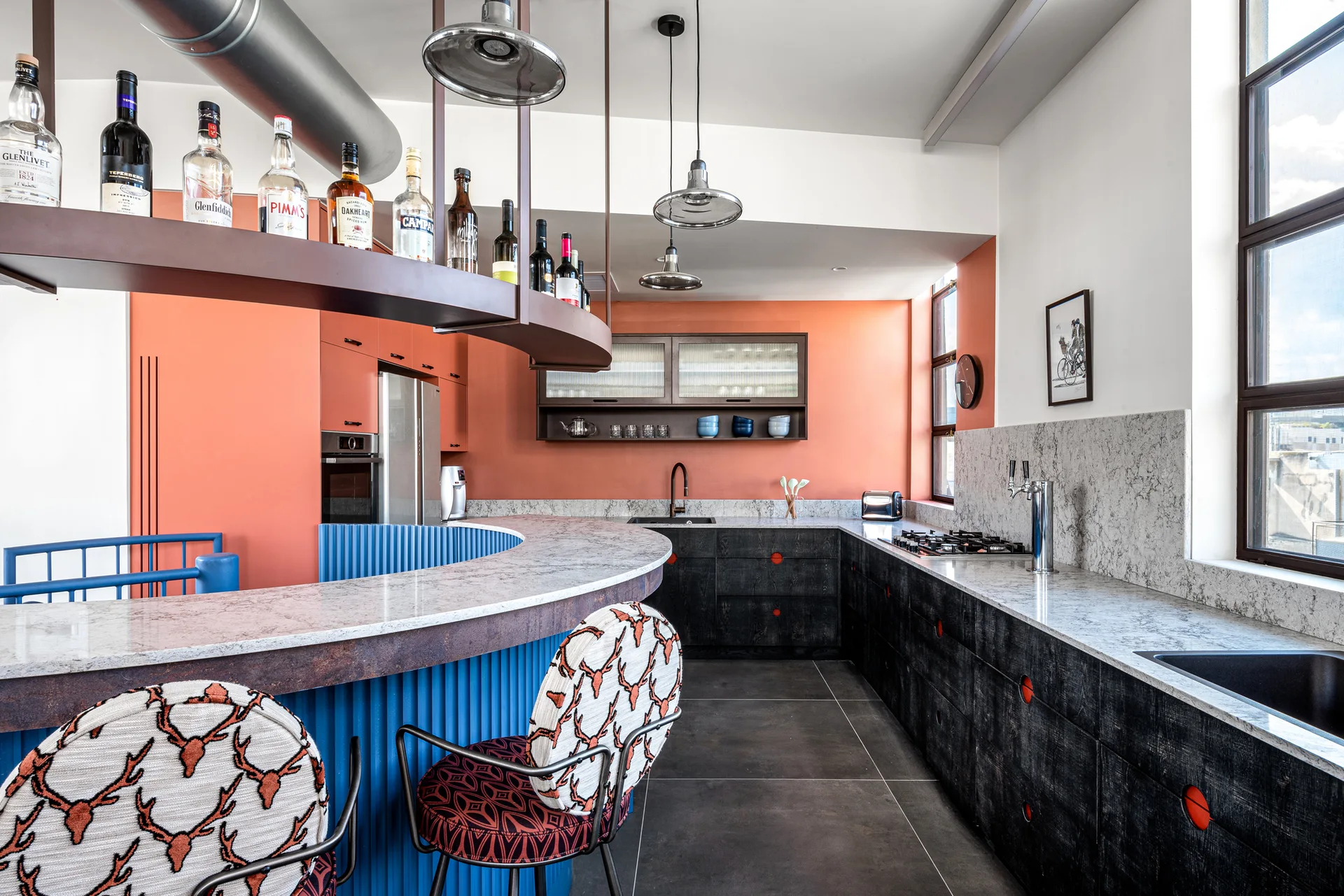 A modern kitchen featuring a curved blue corrugated island, coral walls, black cabinetry, and two patterned bar stools.
