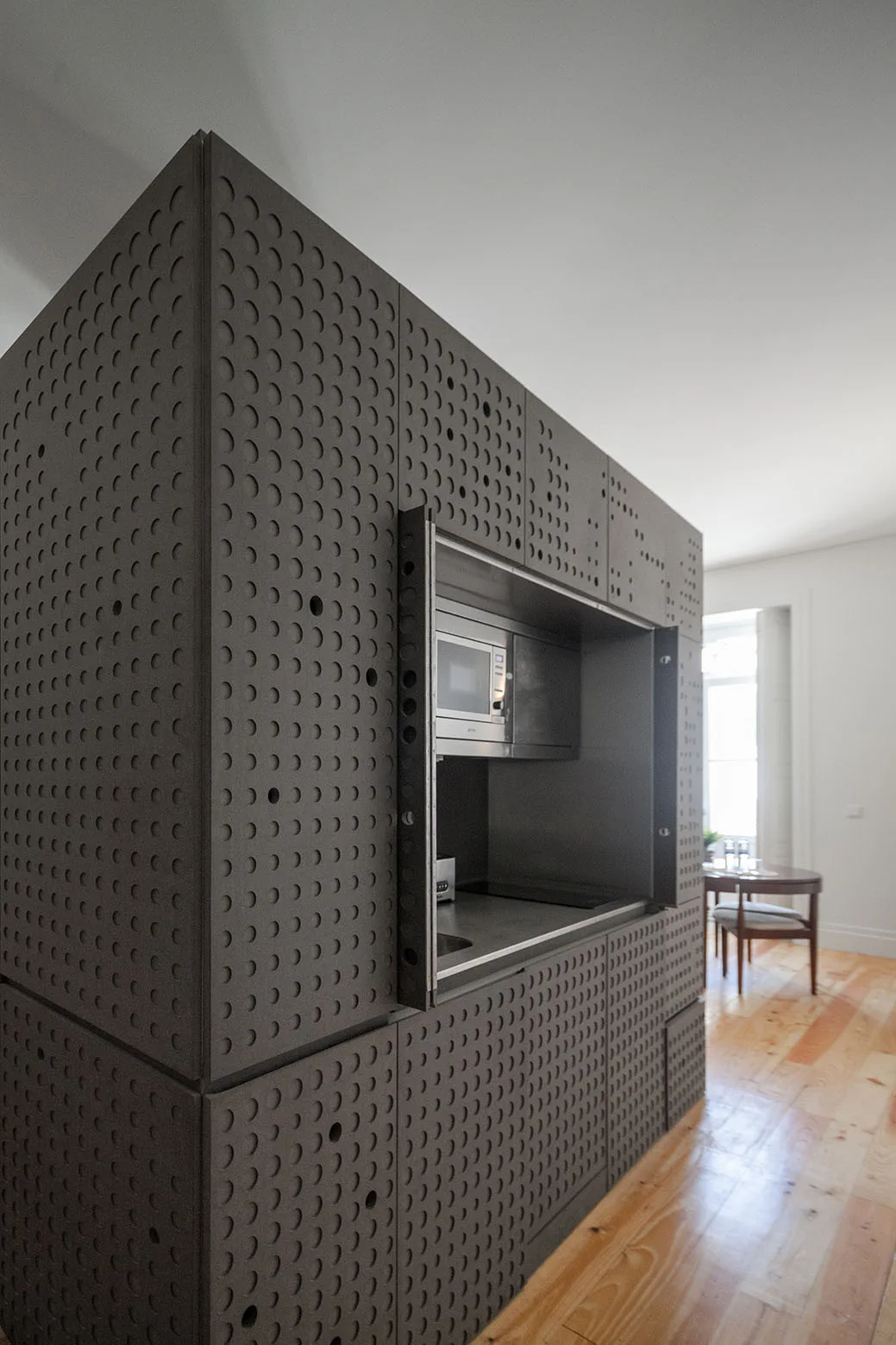 A dark gray monolithic kitchen unit features perforated cabinet doors with circular cutouts and a recessed niche containing a built-in microwave.