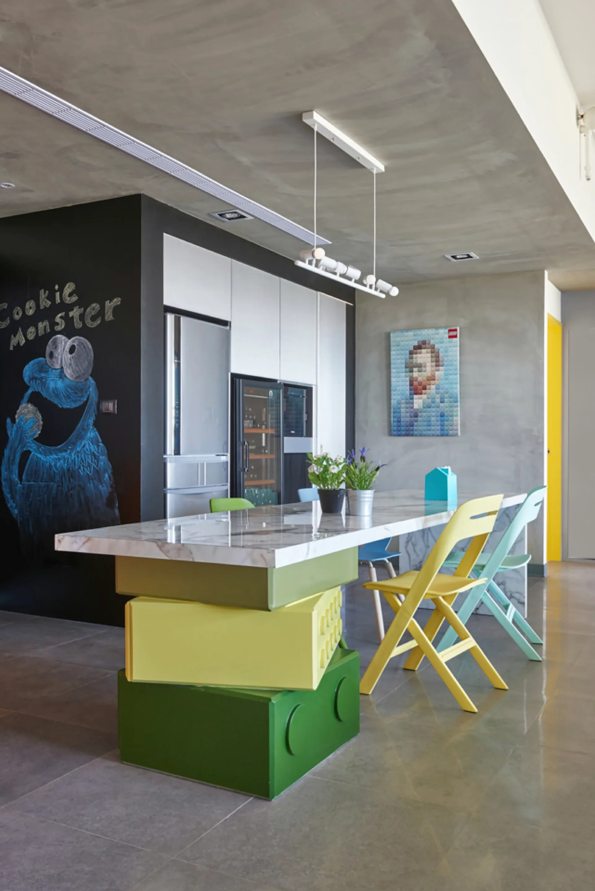 A modern kitchen featuring a marble-topped island with a green building-block base, colorful dining chairs, a concrete ceiling, and a black chalkboard wall.