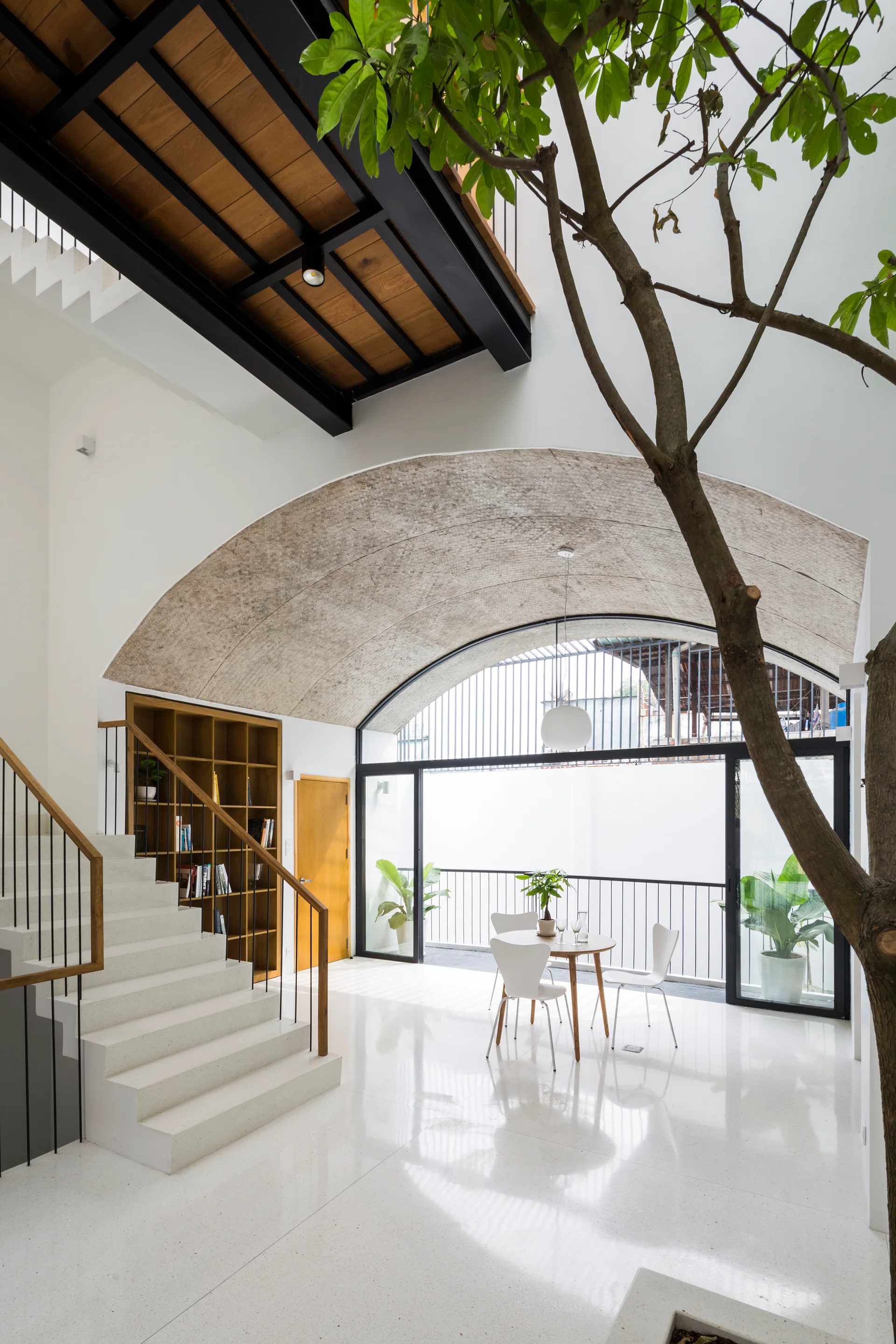 A minimalist home interior with a large concrete vaulted arch, white terrazzo floors, an indoor tree, and a white staircase.