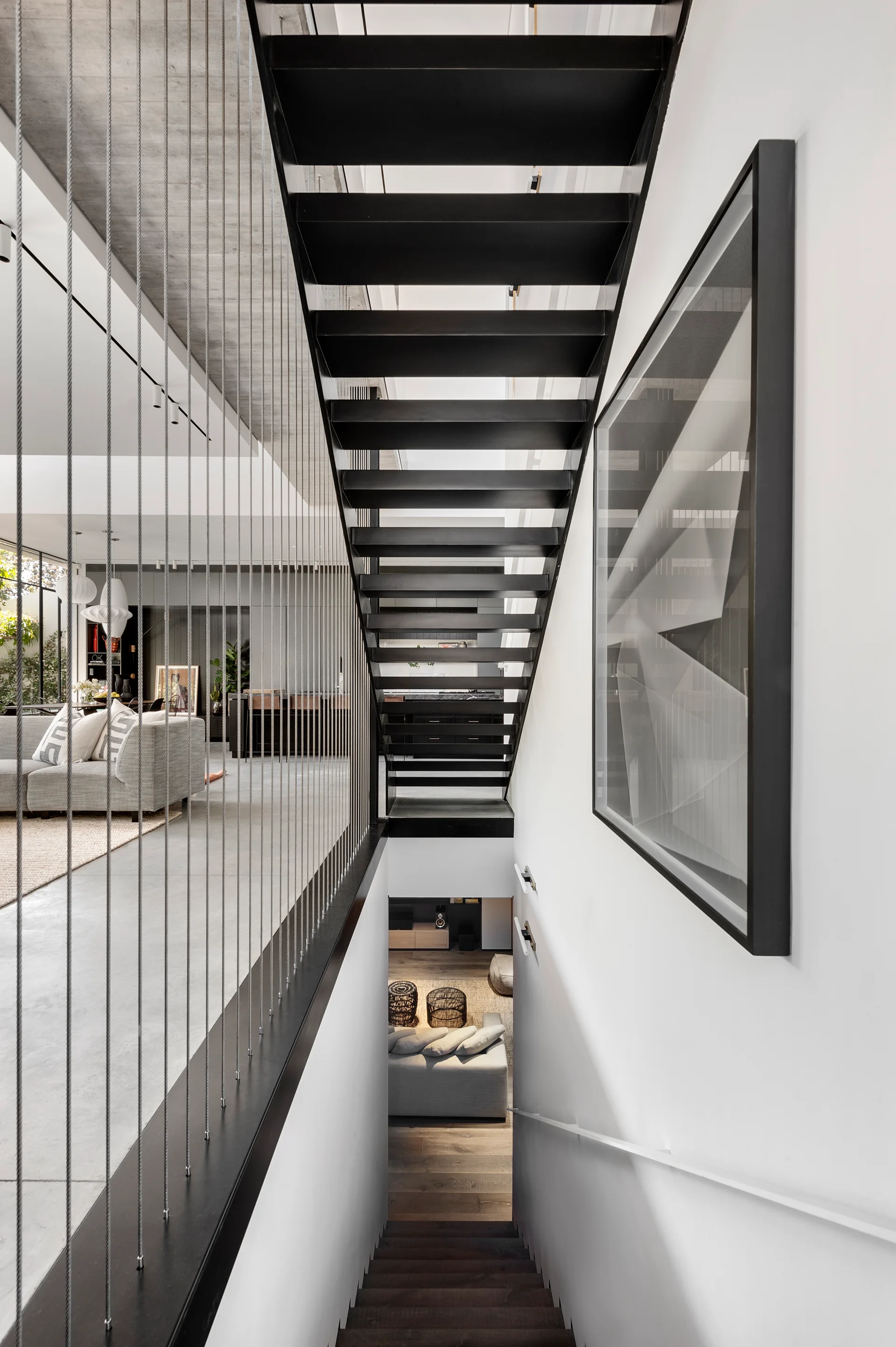 A black metal open-tread staircase features vertical tension cable railings next to smooth white walls and concrete ceilings.