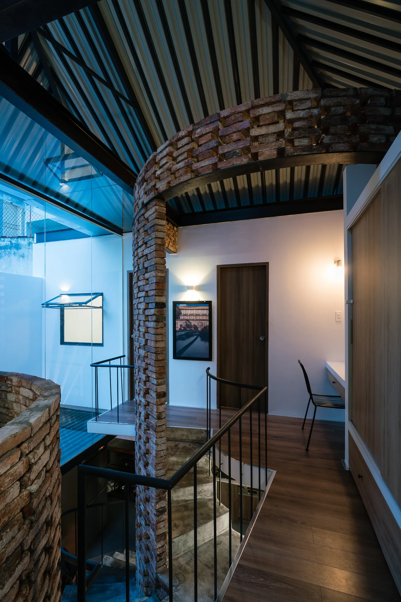 A modern industrial stairwell features a curved exposed brick column, black metal railings, and a vaulted corrugated metal ceiling.