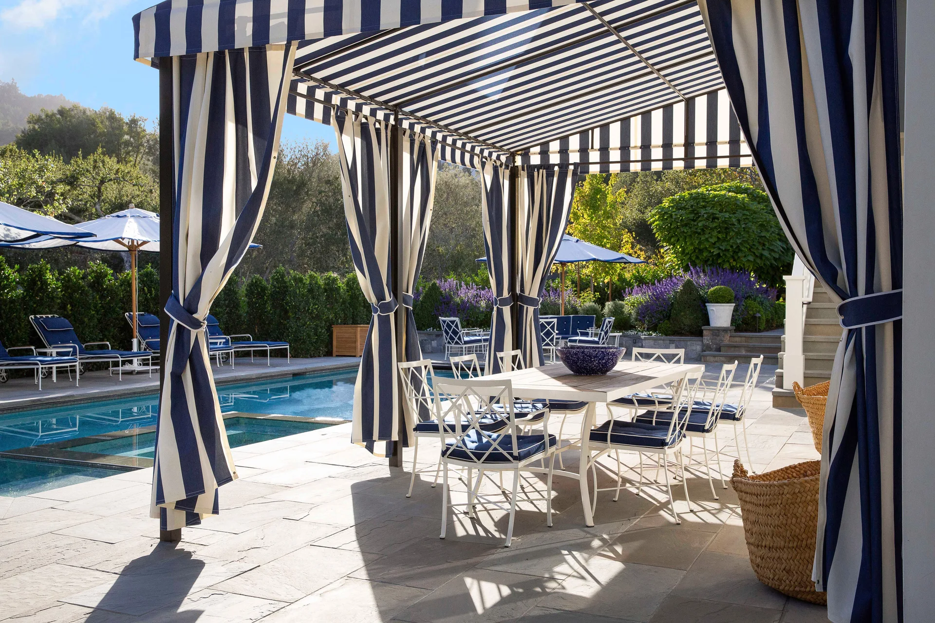 A flagstone poolside patio features a large navy and white striped cabana sheltering a white dining table surrounded by lattice-back chairs.