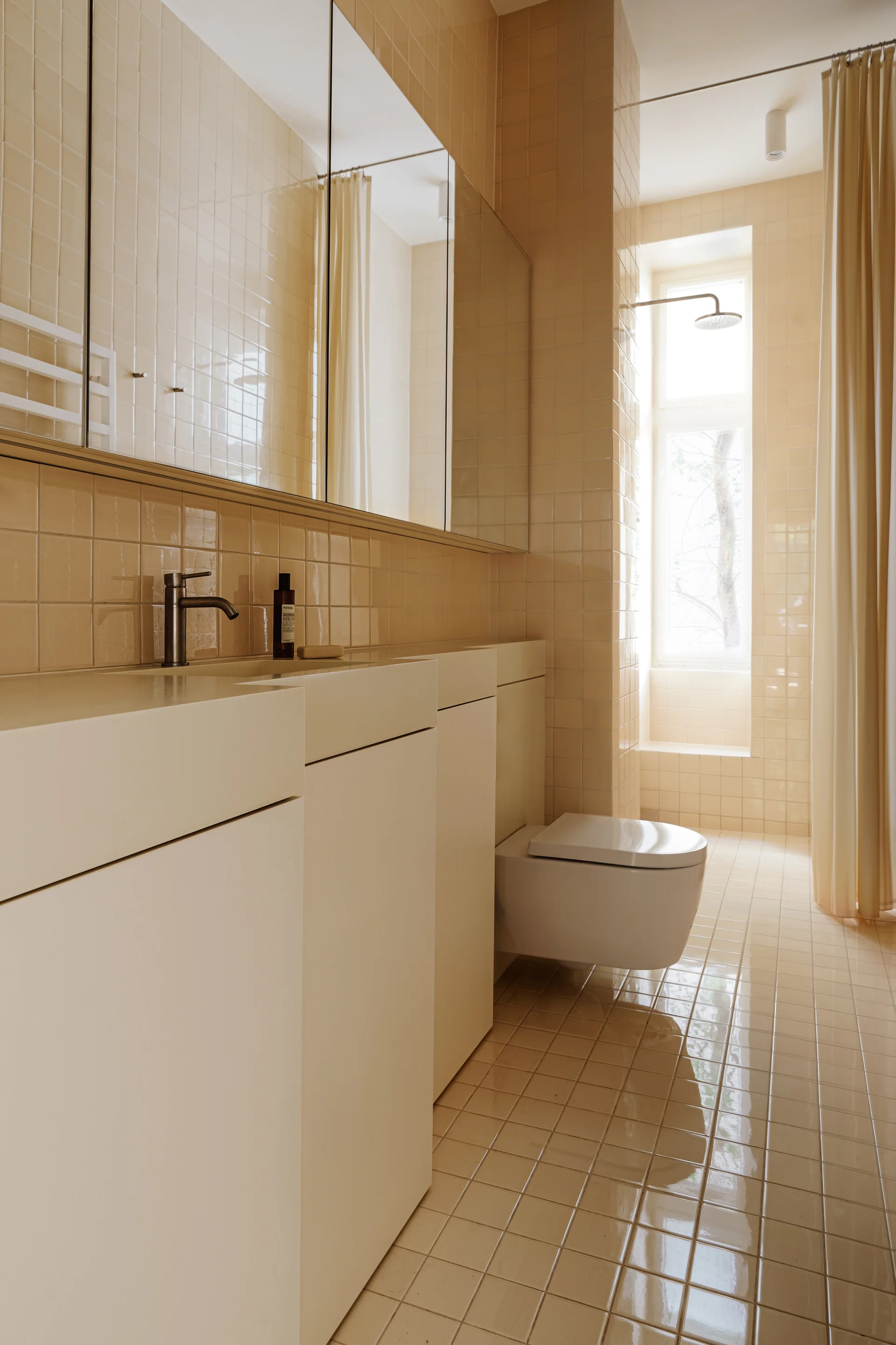 A minimalist bathroom featuring glossy beige square tiles on the floor and walls, a white wall-mounted toilet, and a sleek white vanity.