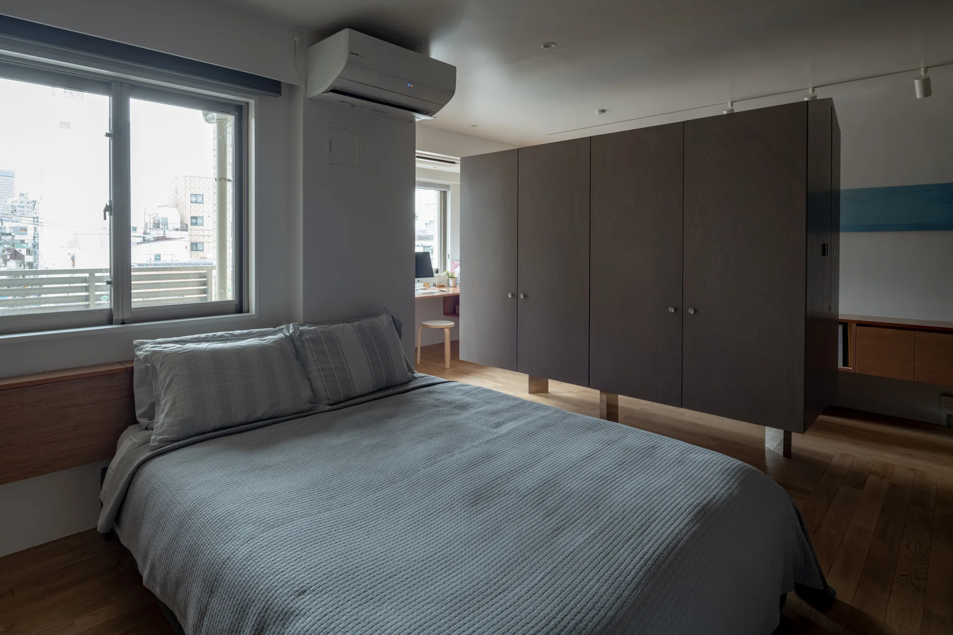 A modern bedroom with grey bedding, wood flooring, and a large freestanding dark wood wardrobe acting as a partition to separate a desk area.