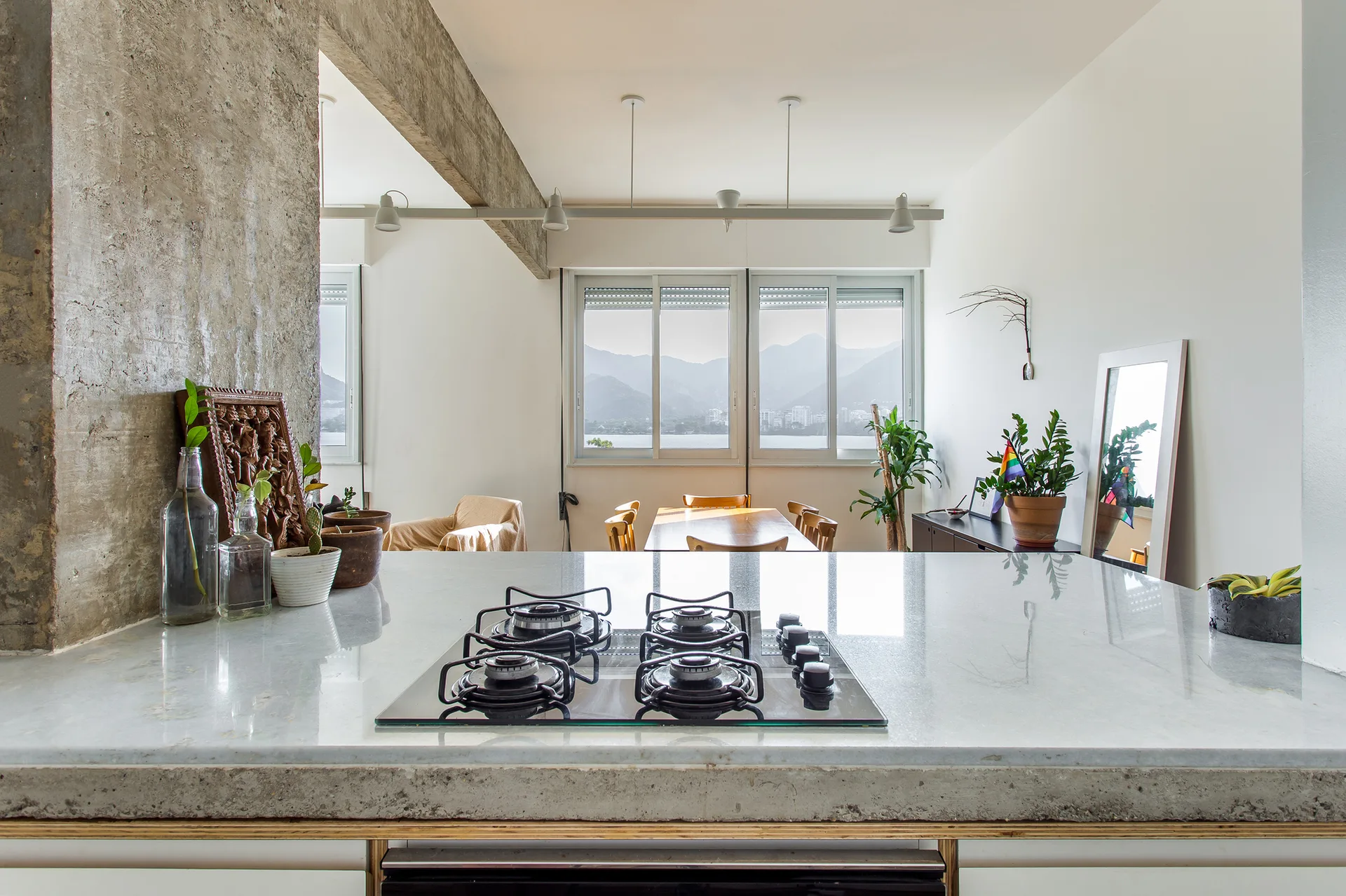 A light-gray kitchen countertop with a gas cooktop overlooks a dining area framed by a raw concrete column and large windows showing mountains.