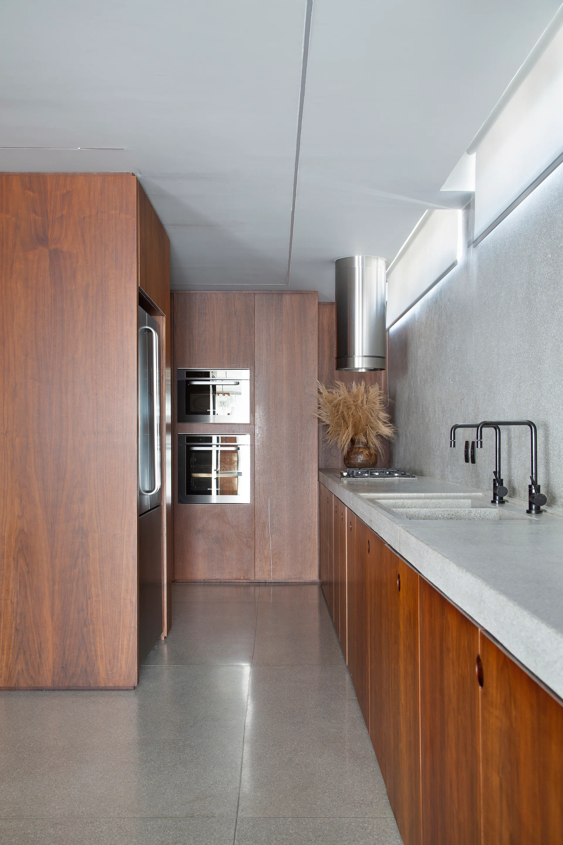 A galley kitchen featuring rich wood-grained cabinetry, a long grey stone countertop with an integrated double sink, black faucets, and steel appliances.