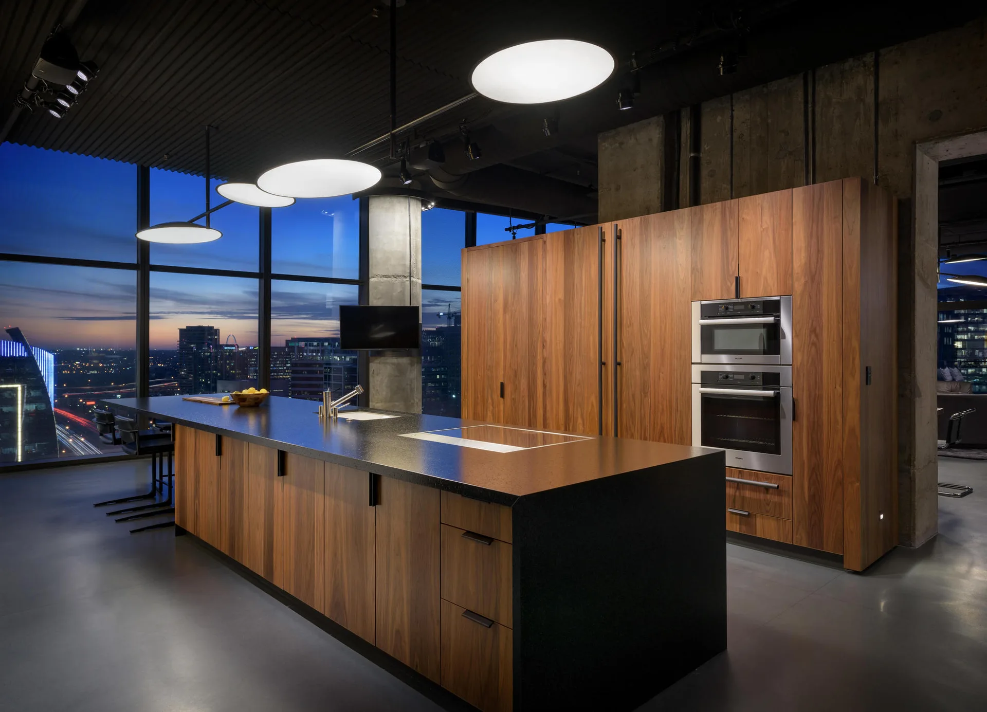 A modern loft kitchen features a dark stone island and matching wood cabinets set against floor-to-ceiling windows with a city night view.