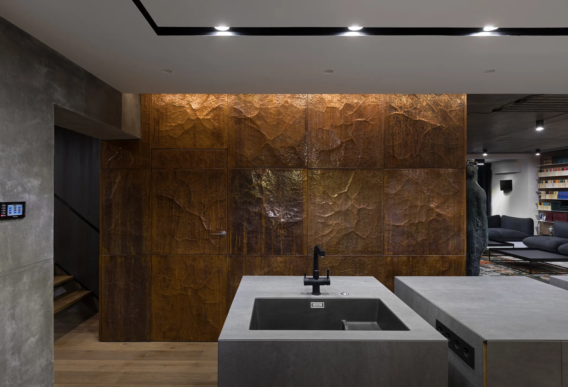A gray stone-look kitchen island with a black sink sits in front of a textured bronze hammered metal feature wall.