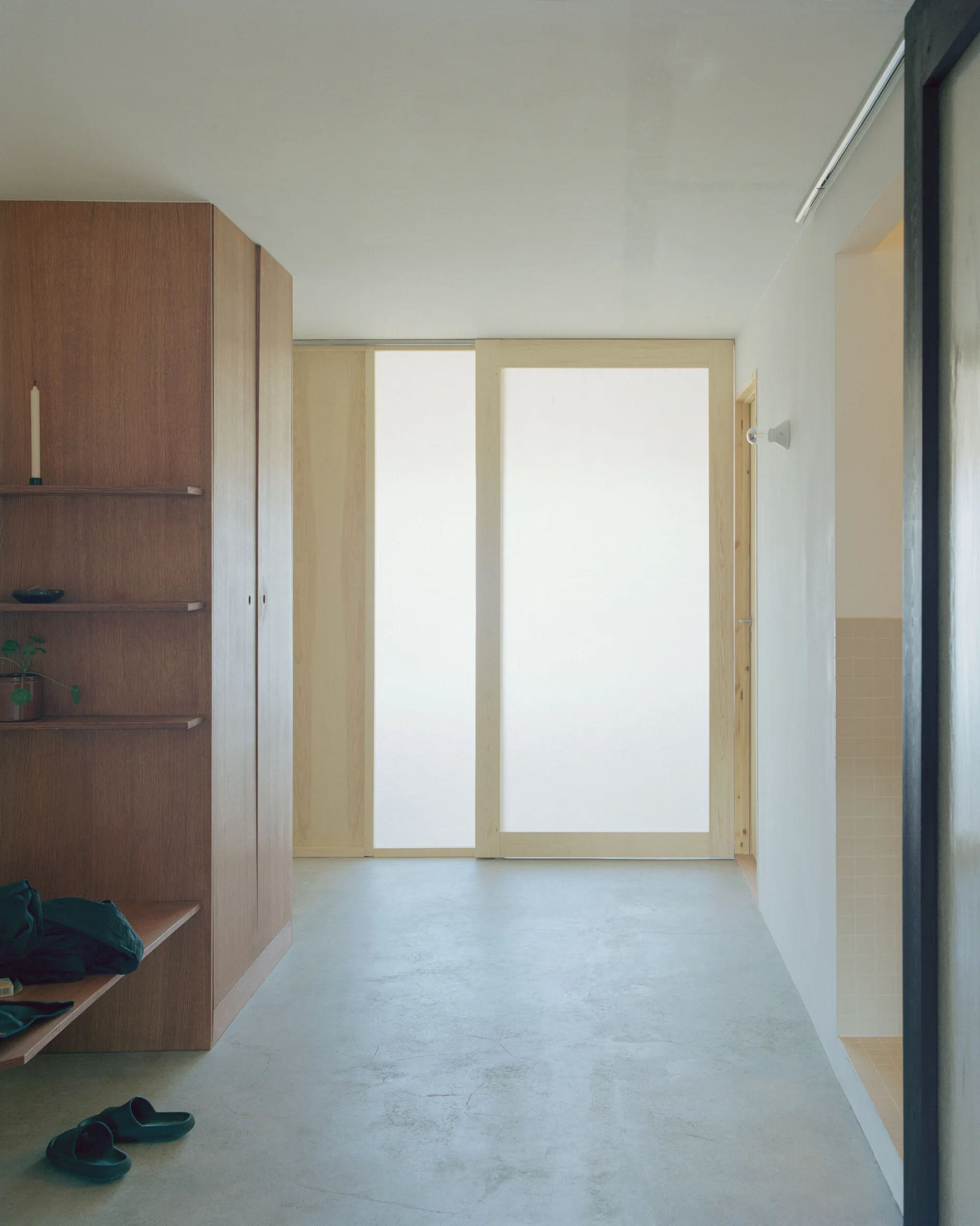 A minimalist entryway features a built-in wooden wardrobe with side shelving, a concrete floor, and frosted glass sliding doors.