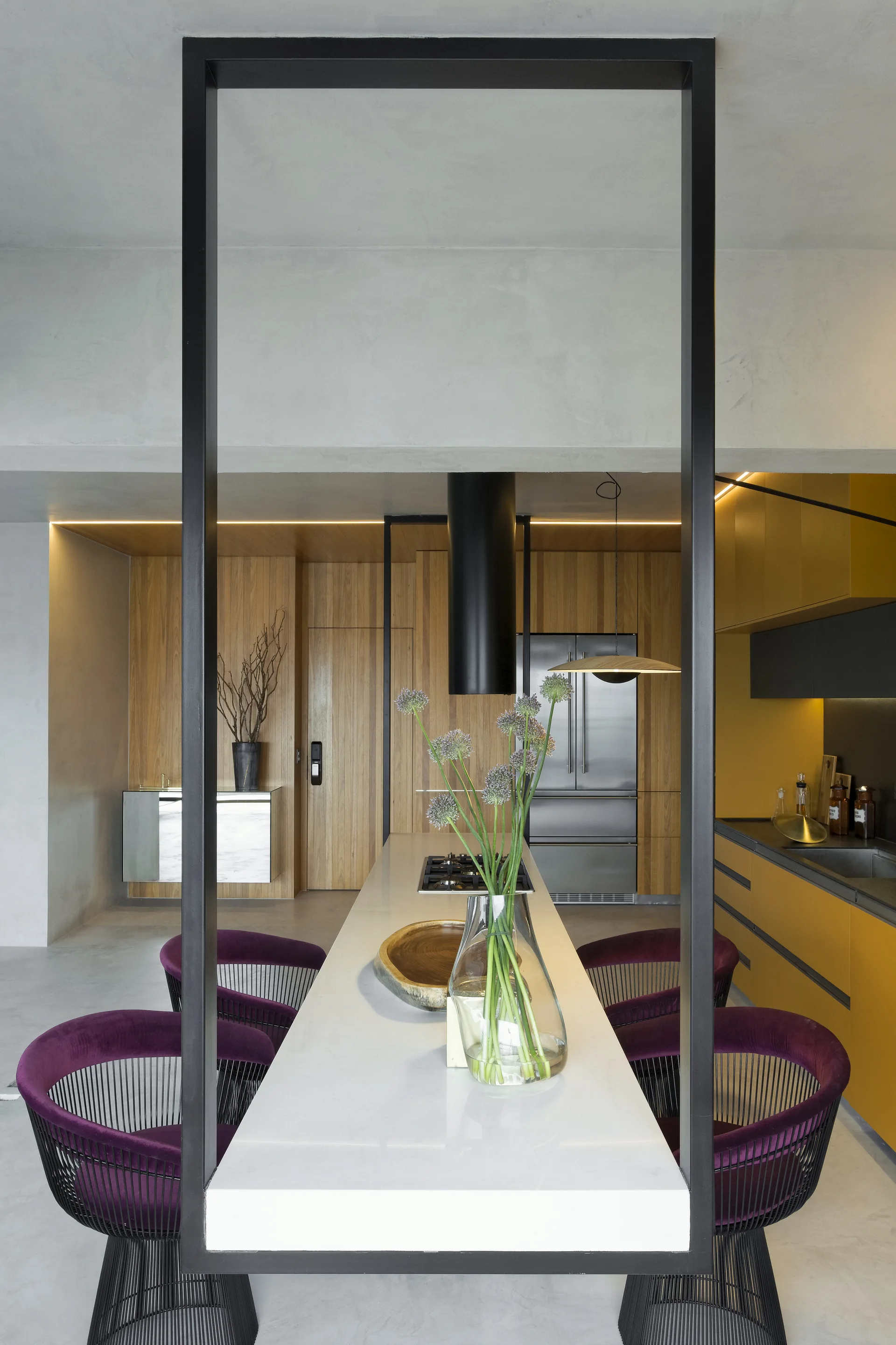 A modern kitchen features a long white island with purple wire-frame chairs, set against mustard yellow cabinets and wood-paneled walls.