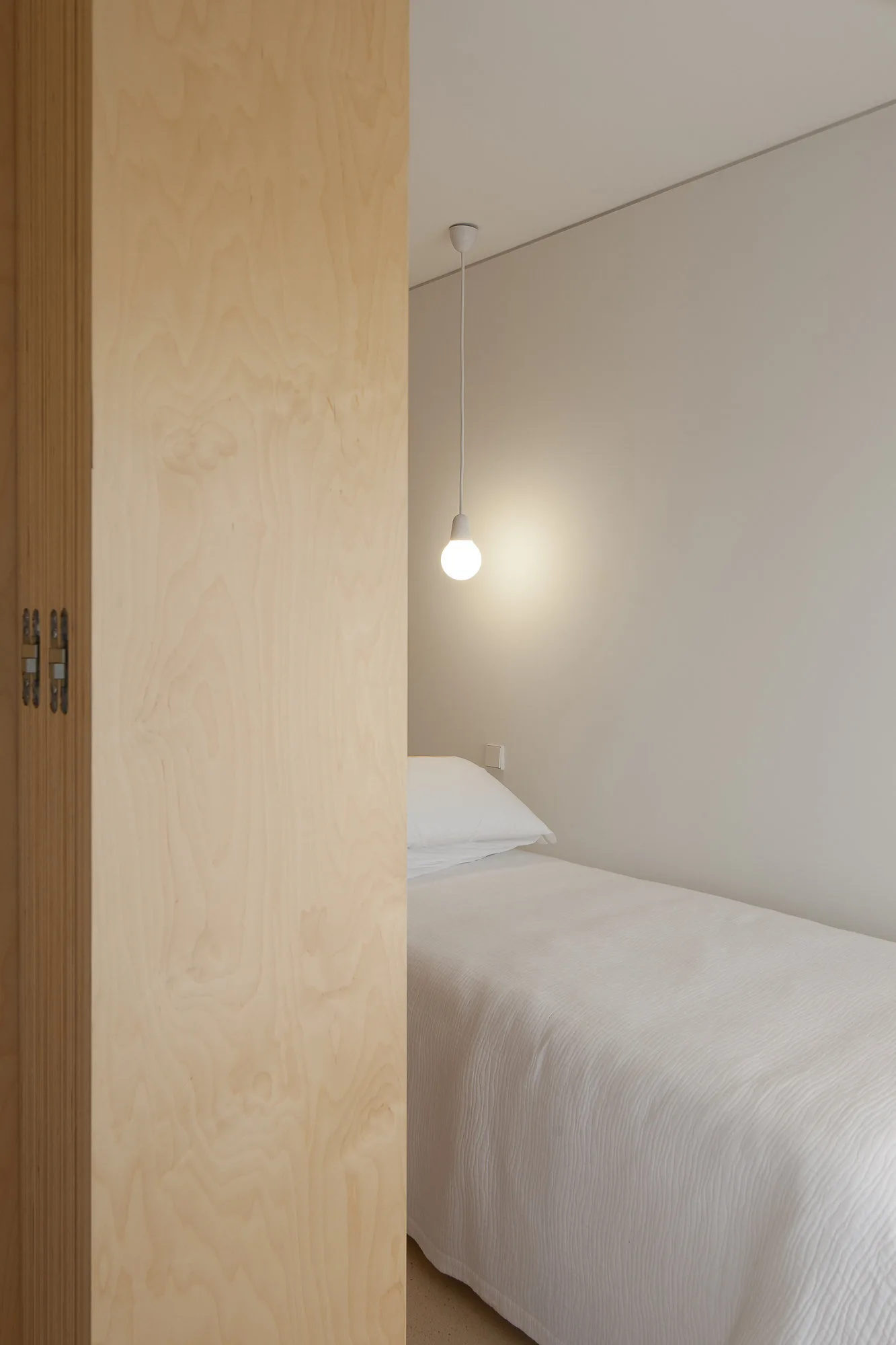 A minimalist bedroom featuring a bed with white bedding, a hanging bulb pendant light, and a light-colored plywood partition wall in the foreground.