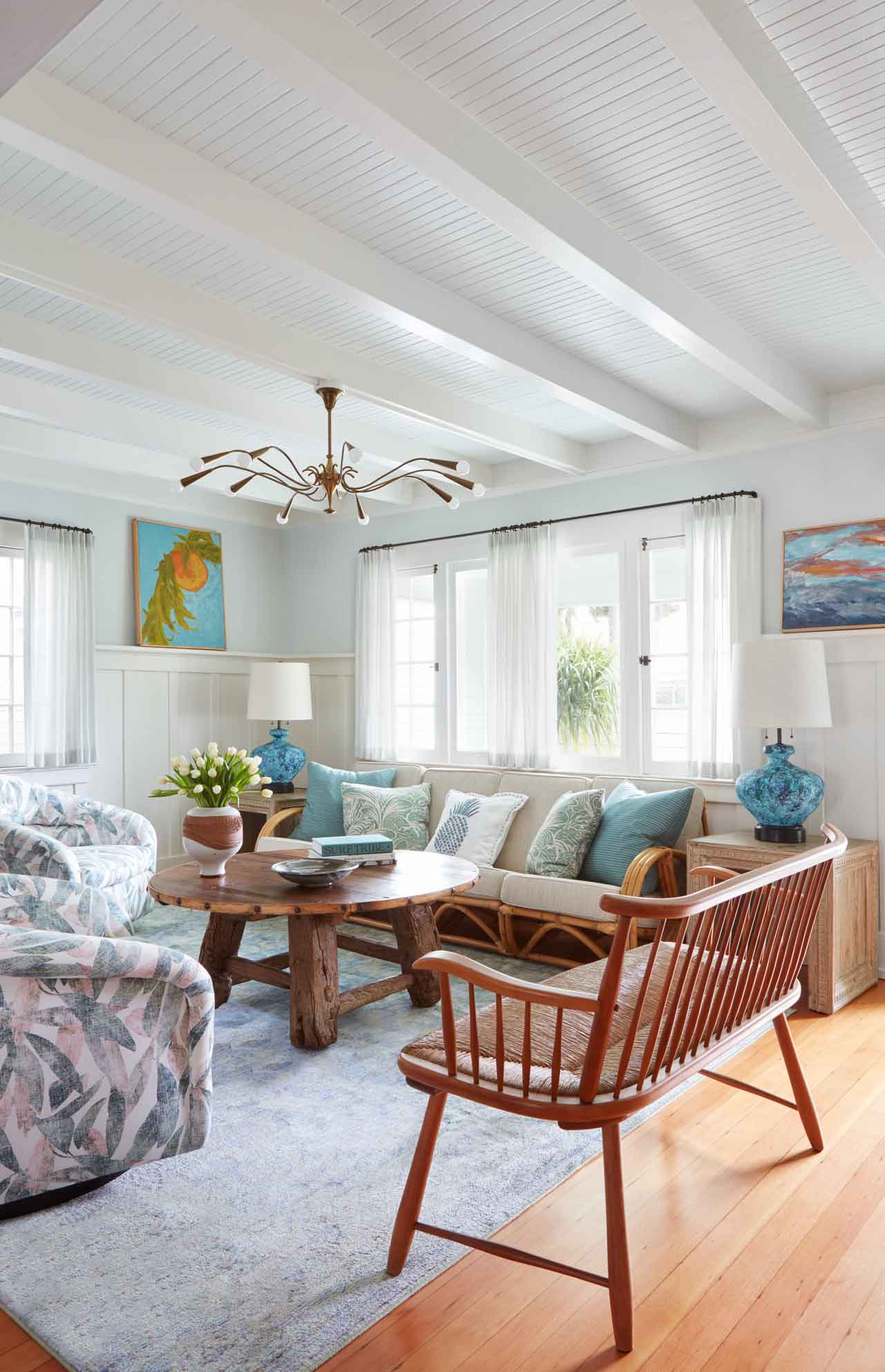 A bright coastal living room featuring a rattan-framed sofa, a round rustic wood coffee table, blue lamps, and white painted ceiling beams.