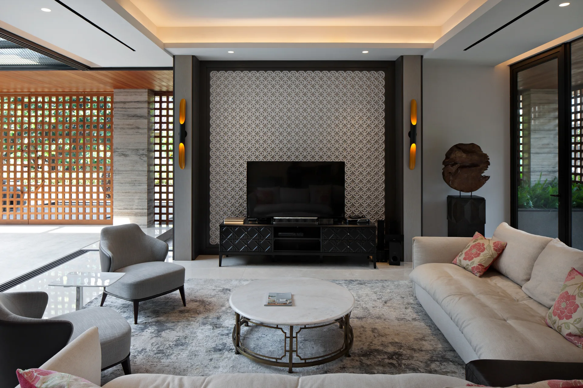 A modern living room features a patterned wall screen behind a television, a cream sofa, and grey armchairs around a marble coffee table.
