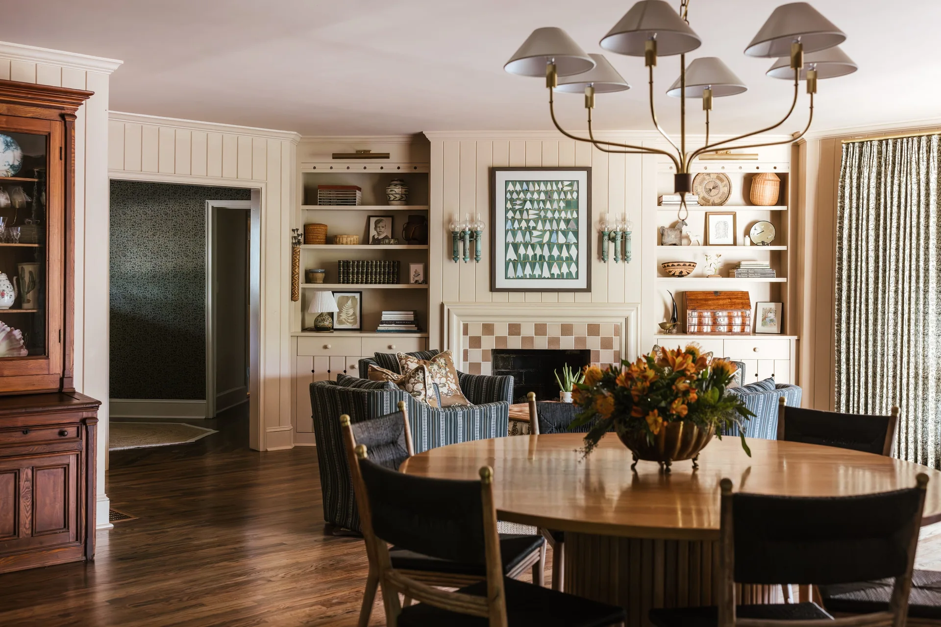 A warm living and dining area features a round wood table in the foreground, striped armchairs, and built-in bookshelves flanking a tiled fireplace.