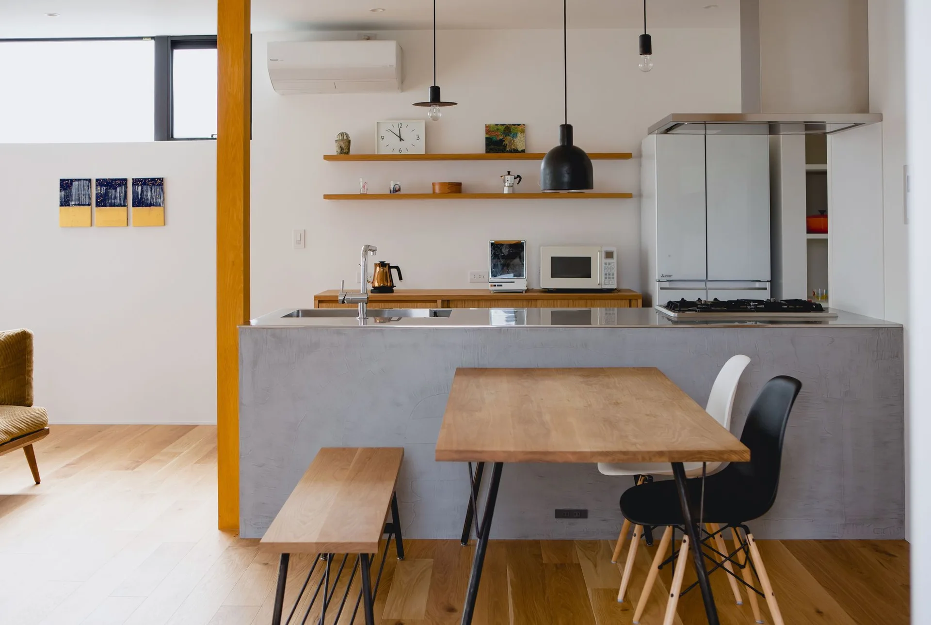 A minimalist open kitchen features a grey concrete island, a wooden dining table with a matching bench, and light wood flooring under white walls.