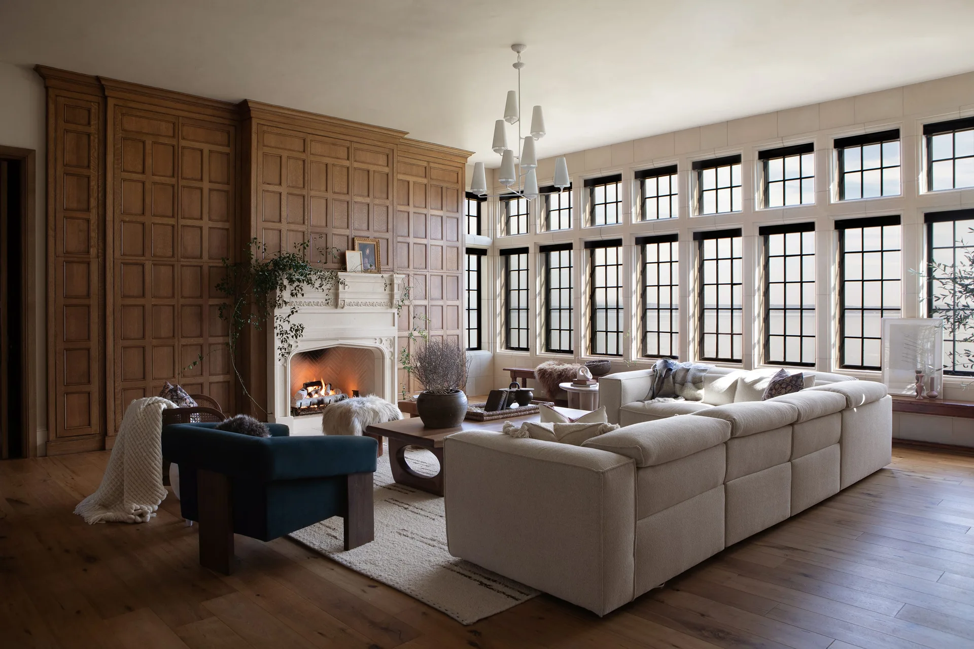 A spacious living room features extensive wood-paneled walls, a stone fireplace, a cream sectional, and a wall of black grid windows.