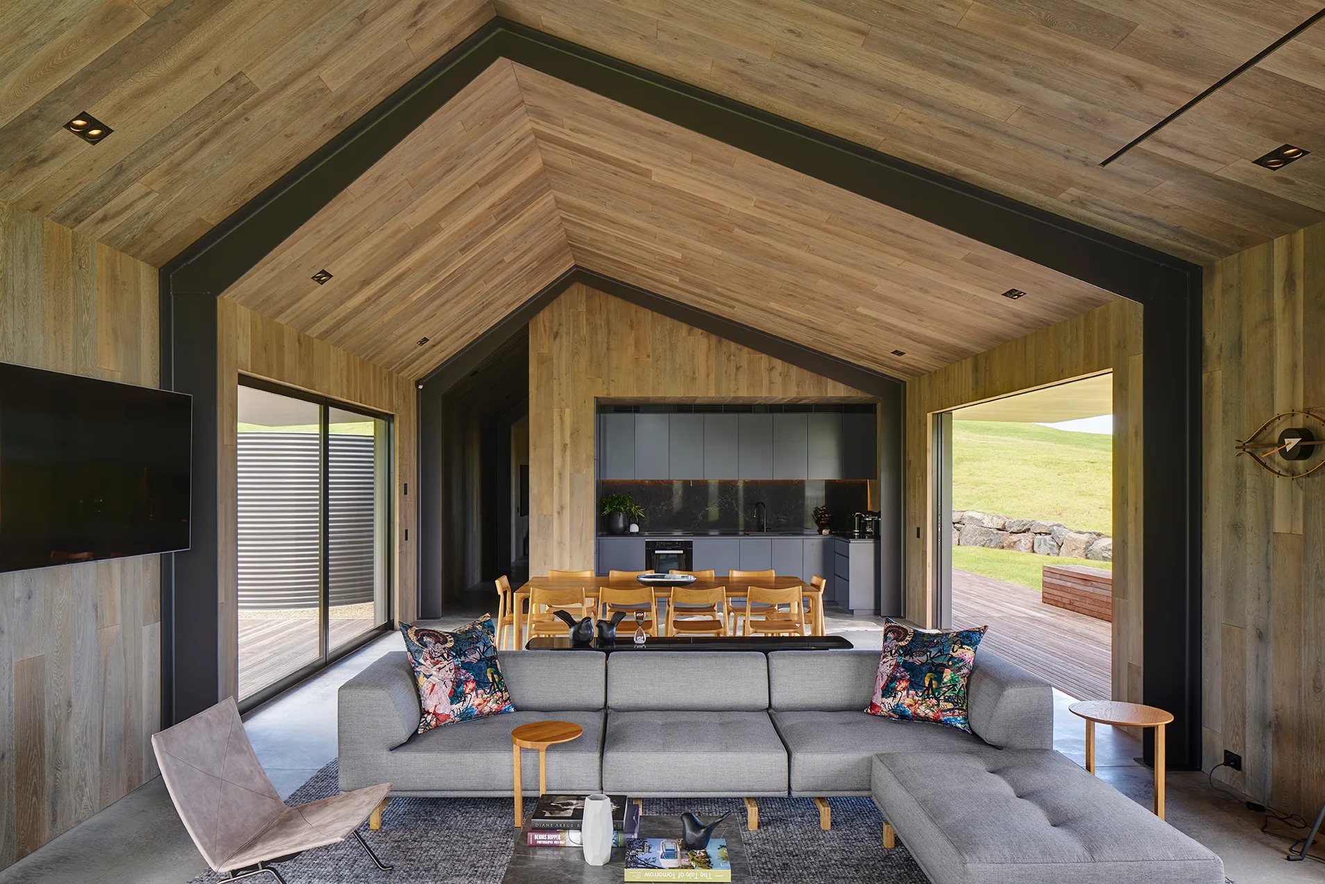 A modern open-concept living room features a grey sectional sofa, vaulted timber-clad ceilings with black steel framing, and sliding glass doors.