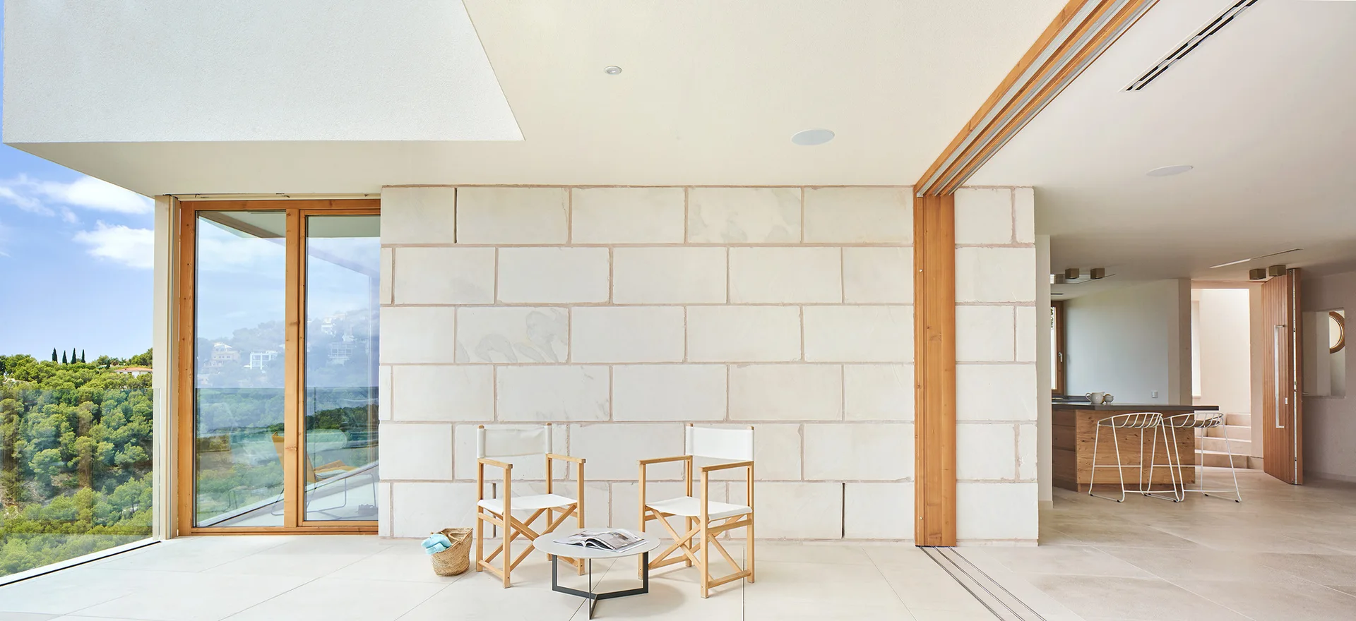 A minimalist covered patio features two wood and white canvas director chairs, a small round table, and a large-block cream stone feature wall.