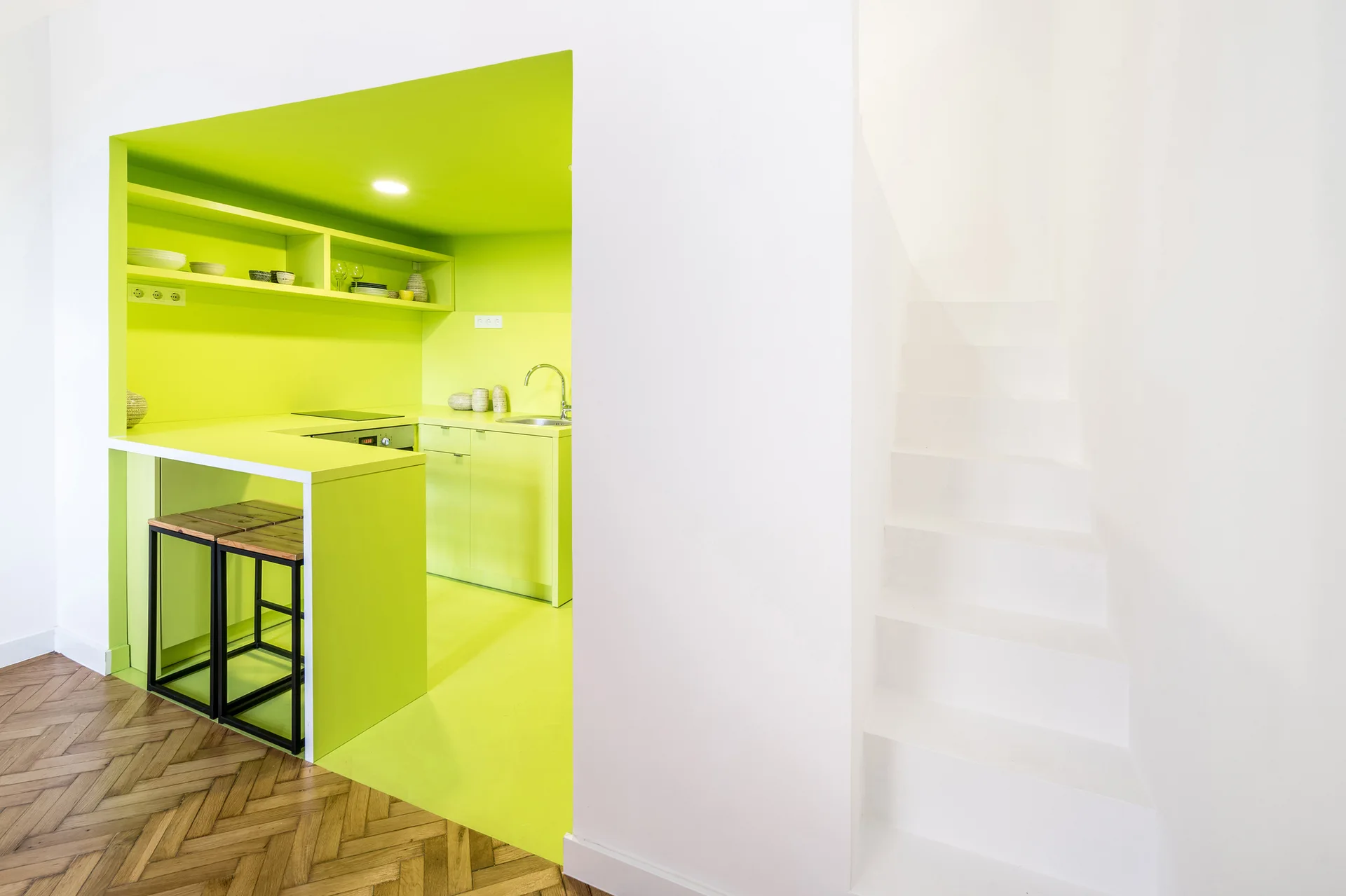 A bright lime green recessed kitchen features matching cabinetry and stools next to a minimalist white staircase and herringbone wood floors.